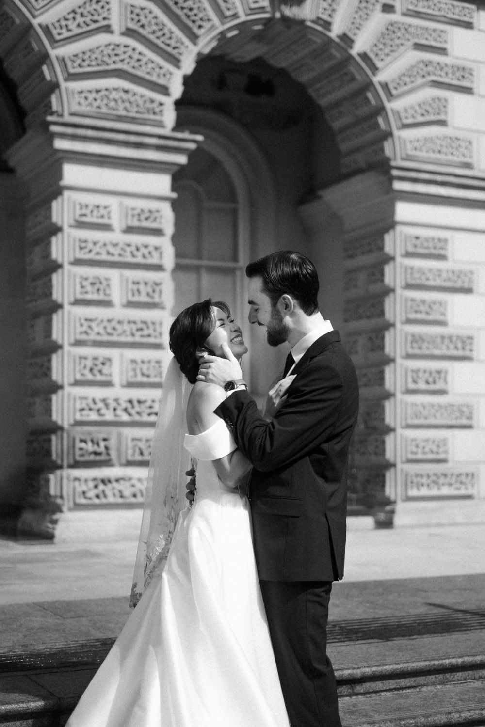 Black and white photo of a bride and groom embracing before an ornate arched building on their wedding day.