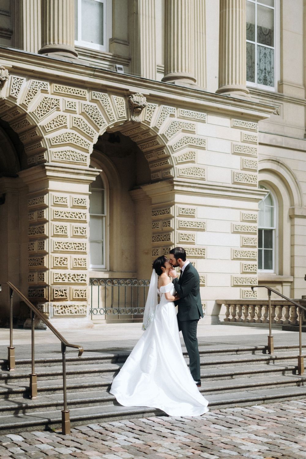 Bride and groom share a romantic moment on cobblestone steps outside an elegant neoclassical building.