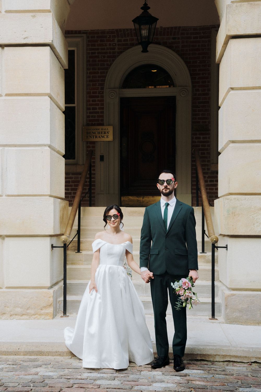 Bride and groom in sunglasses hold hands on steps of historic brick building, groom in dark green suit.