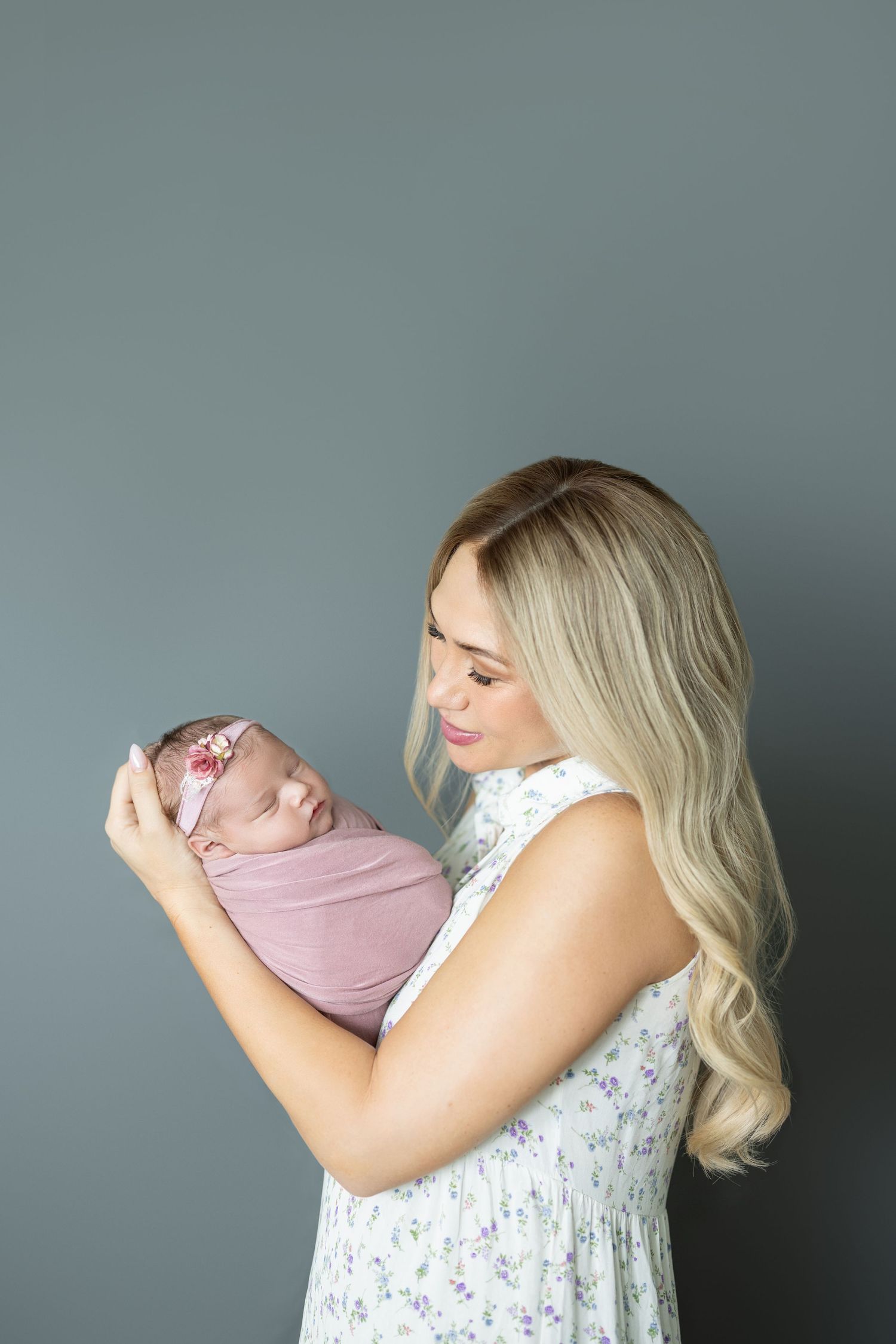 Blonde mother in floral dress tenderly holds swaddled newborn baby girl with pink headband against gray background.
