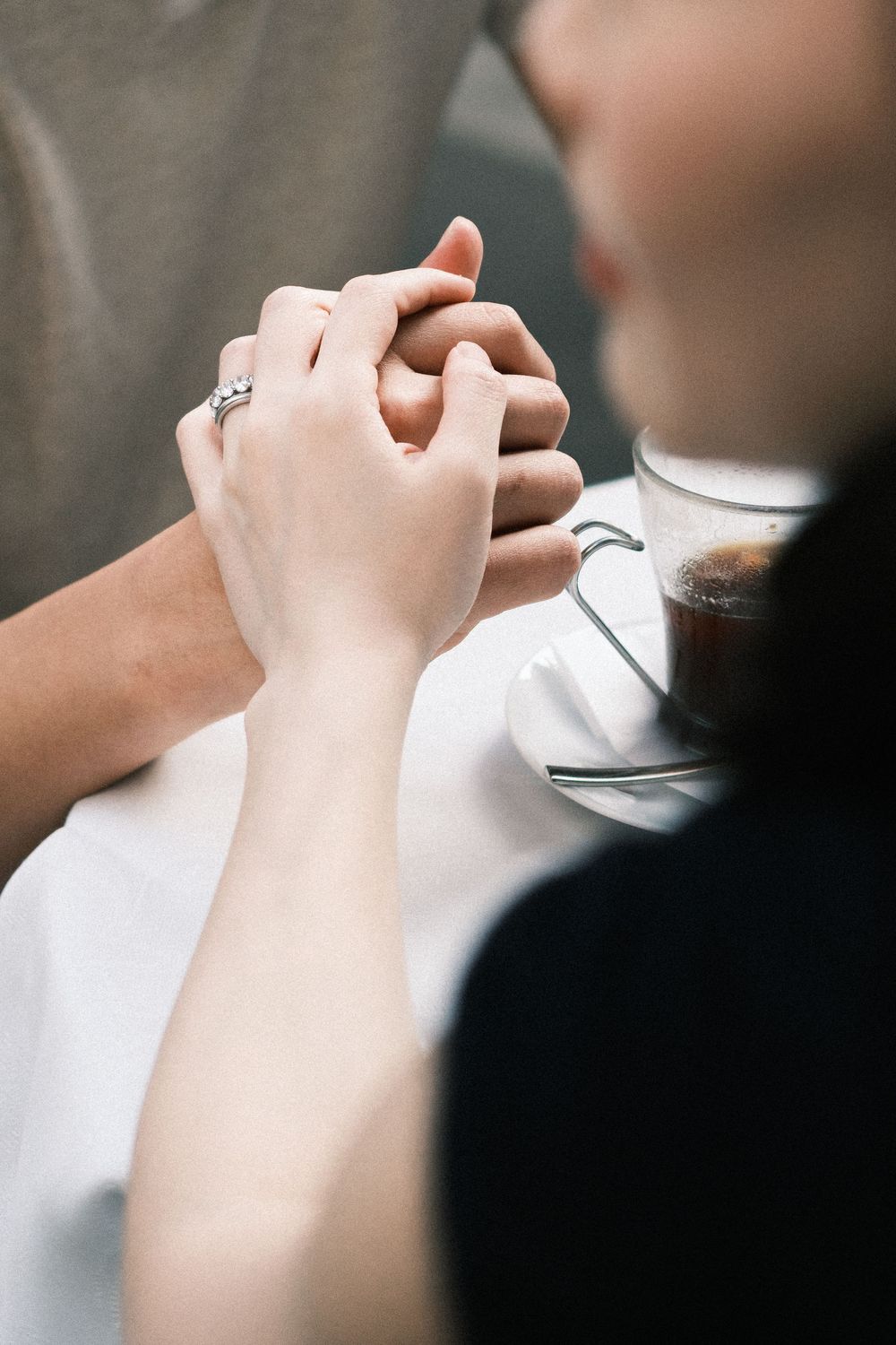 Couple holding hands tenderly over a coffee cup, woman wearing a diamond ring, romantic cafe moment.