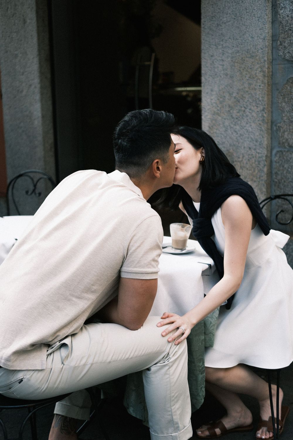 Couple sharing a romantic kiss at an outdoor cafe table with coffee, dressed in casual white and black outfits.