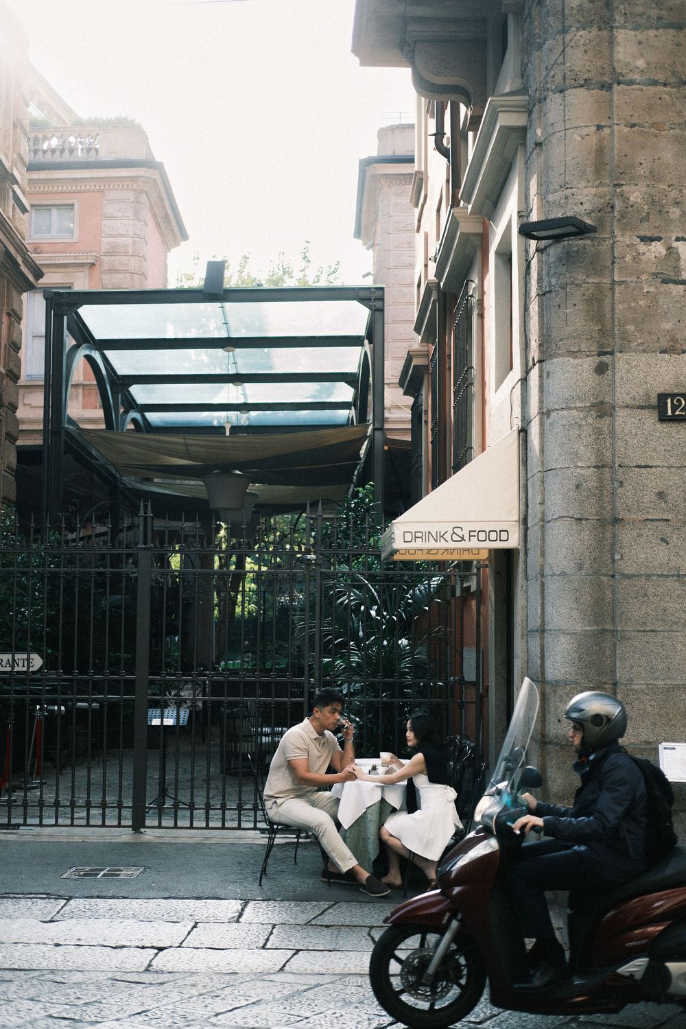 Couple dining at outdoor cafe terrace beside a scooter on a cobblestone street near a Drink & Food sign.