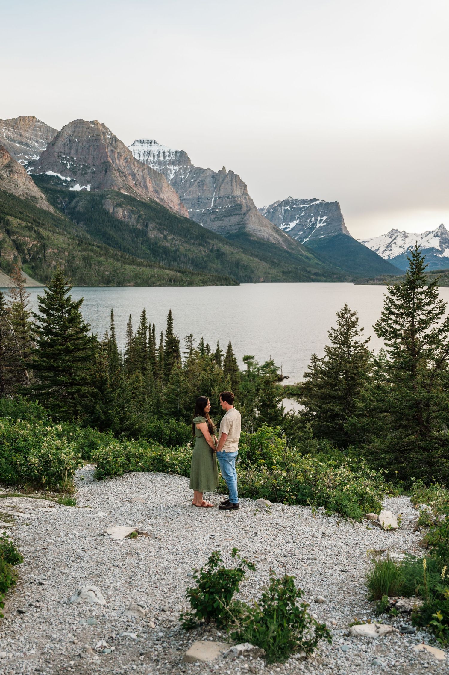 Couple holds hands on rocky overlook with Saint Mary Lake and snow-capped Glacier National Park mountains behind them.
