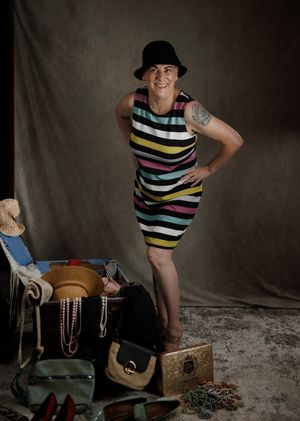 A smiling person in a colorful striped dress and black fedora poses confidently beside vintage steamer trunk and accessories.
