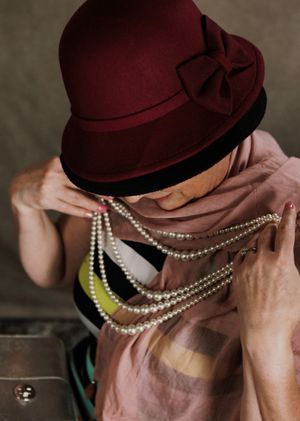 Woman wearing a burgundy cloche hat with bow, and pink scarf, looking downward while holding up a mulit strand pearl costume necklace.