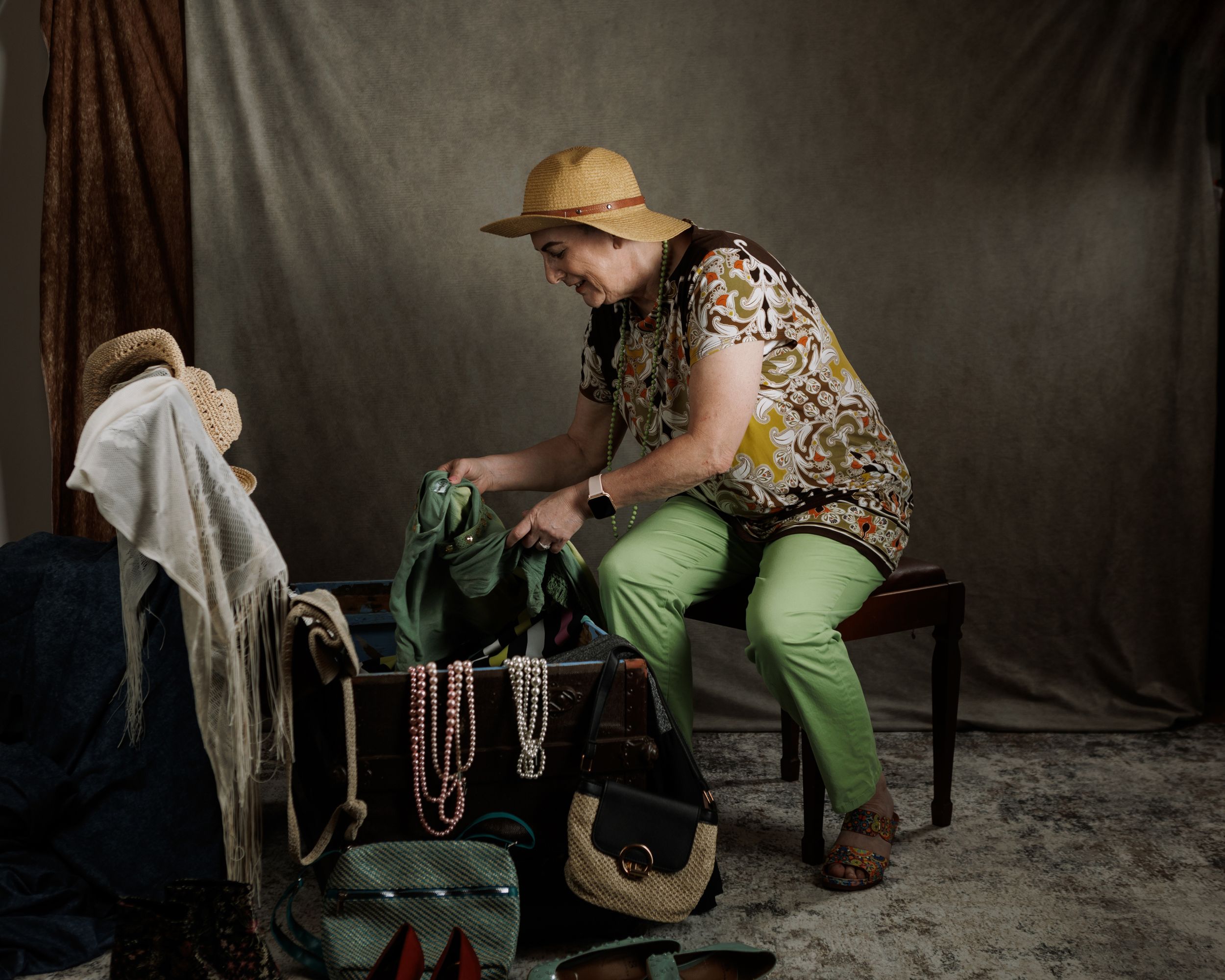 Woman in yellow hat and green pants sorting through an old trunk full of  vintage clothes and jewelry.