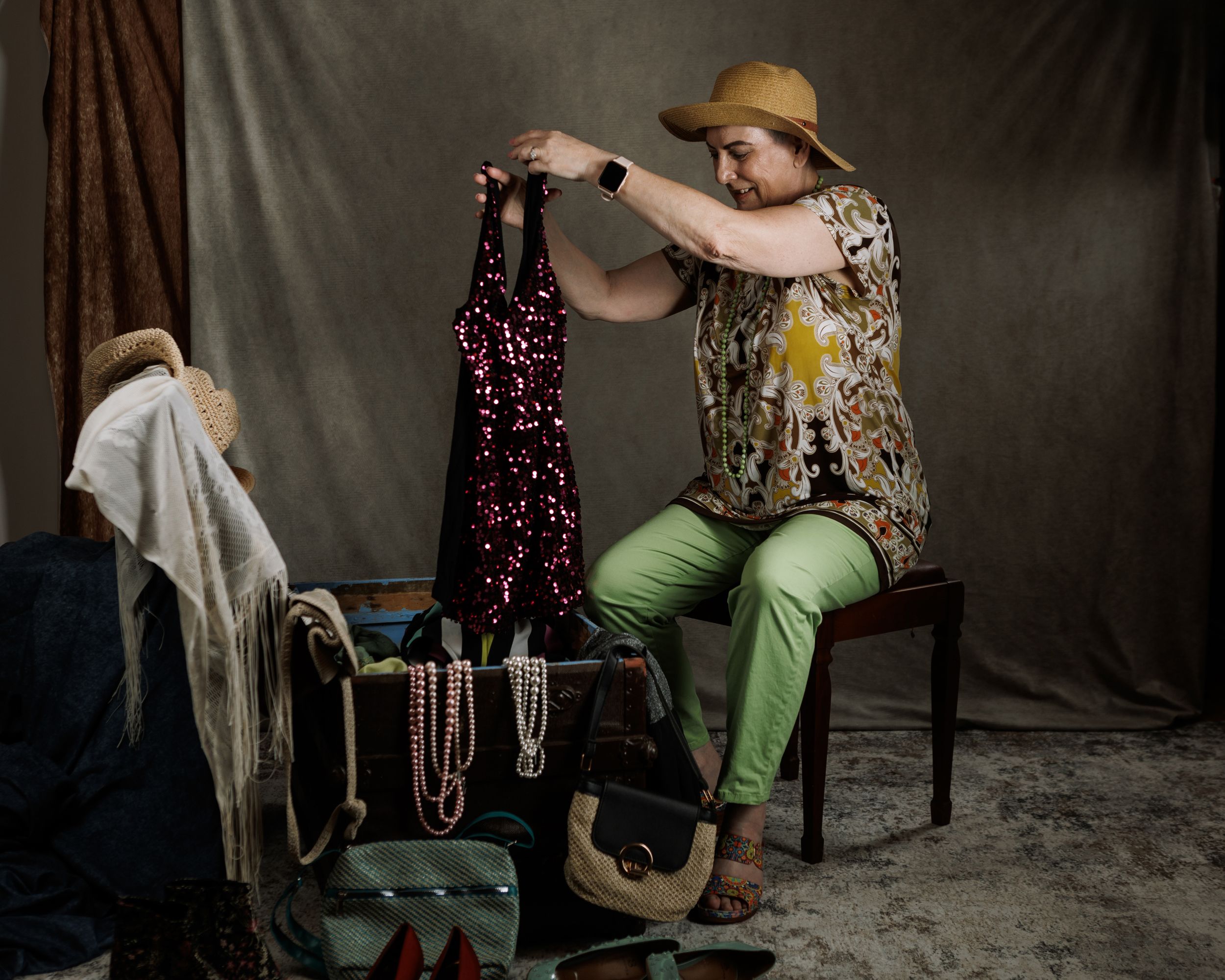 Woman in green pants and floral top examines sequined dress while seated among vintage accessories and jewelry.