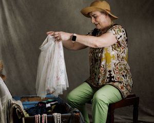 Woman in yellow hat and green pants sorting through an old trunk examining a white lace garment