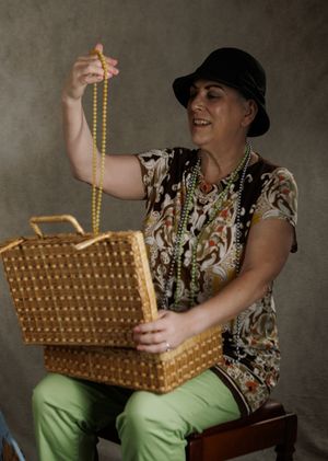 Woman in black hat holds yellow beaded necklace from vintage wicker basket, wearing green pants and floral top.