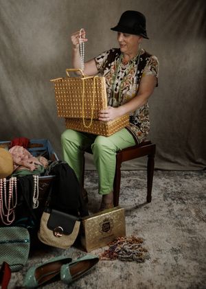 Woman in black hat and green pants holding pearl necklace from gold basket, surrounded by bags and jewelry on floor.