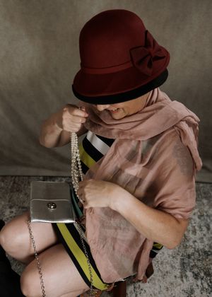 Woman wearing a burgundy cloche hat with bow, and pink scarf and clutch bag while holding up a mulit strand pearl costume necklace.