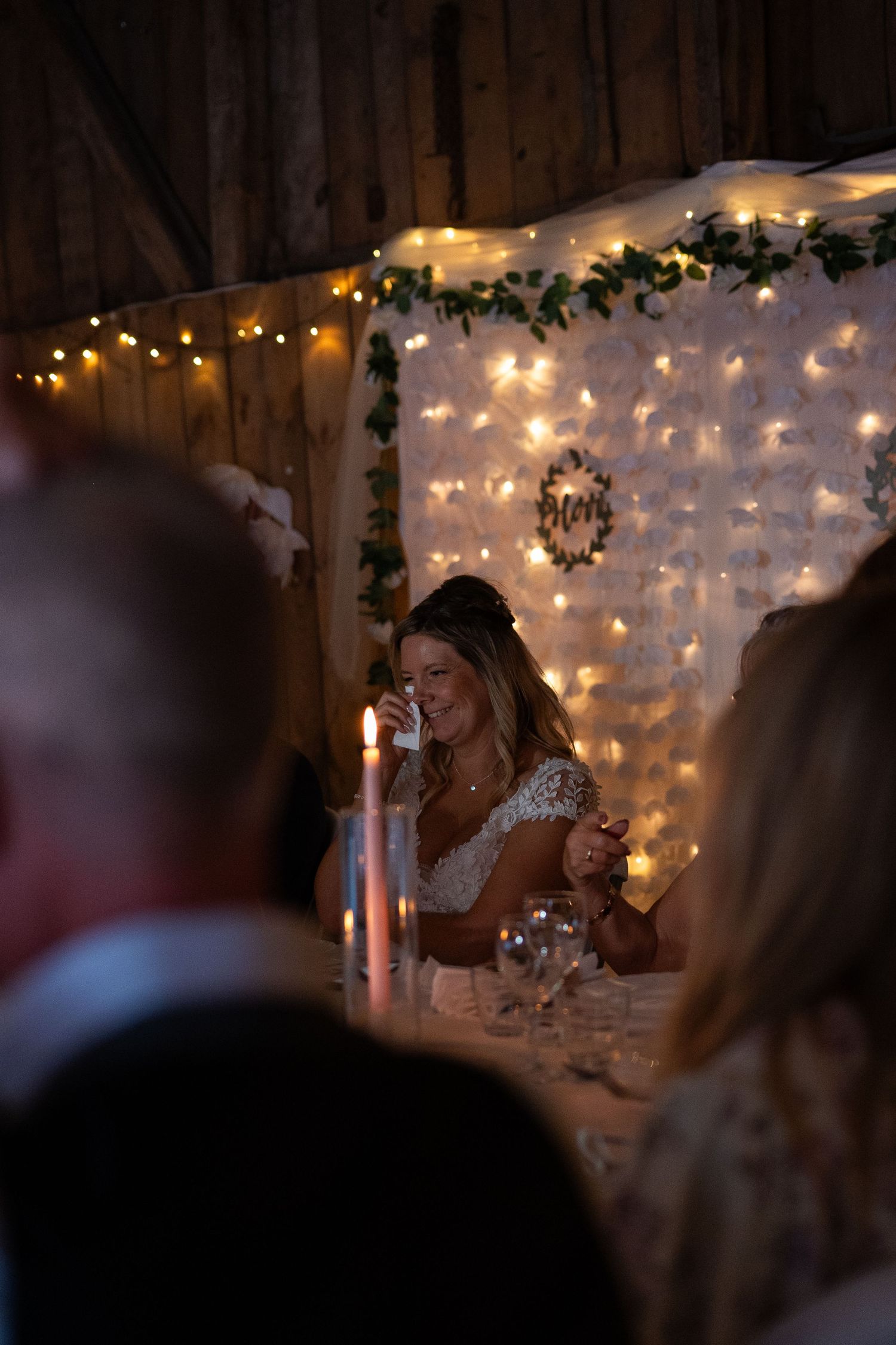 Bride smiling at candlelit wedding reception table with fairy lights and greenery backdrop in rustic barn venue.