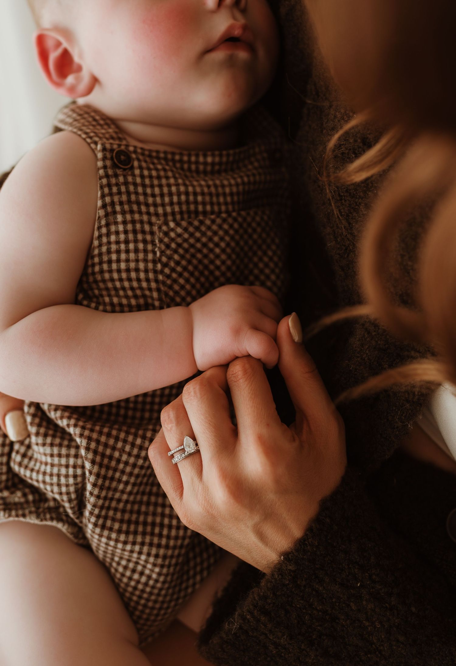 Baby in white dress plays peek-a-boo under cream blanket on textured bedding in warm lighting.