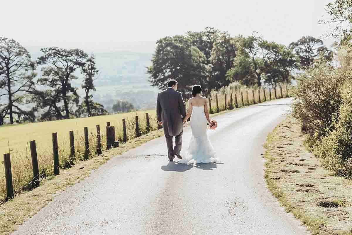 A bride and groom walk hand-in-hand down the path leading from Roundthorn Country House Hotel near Penrith