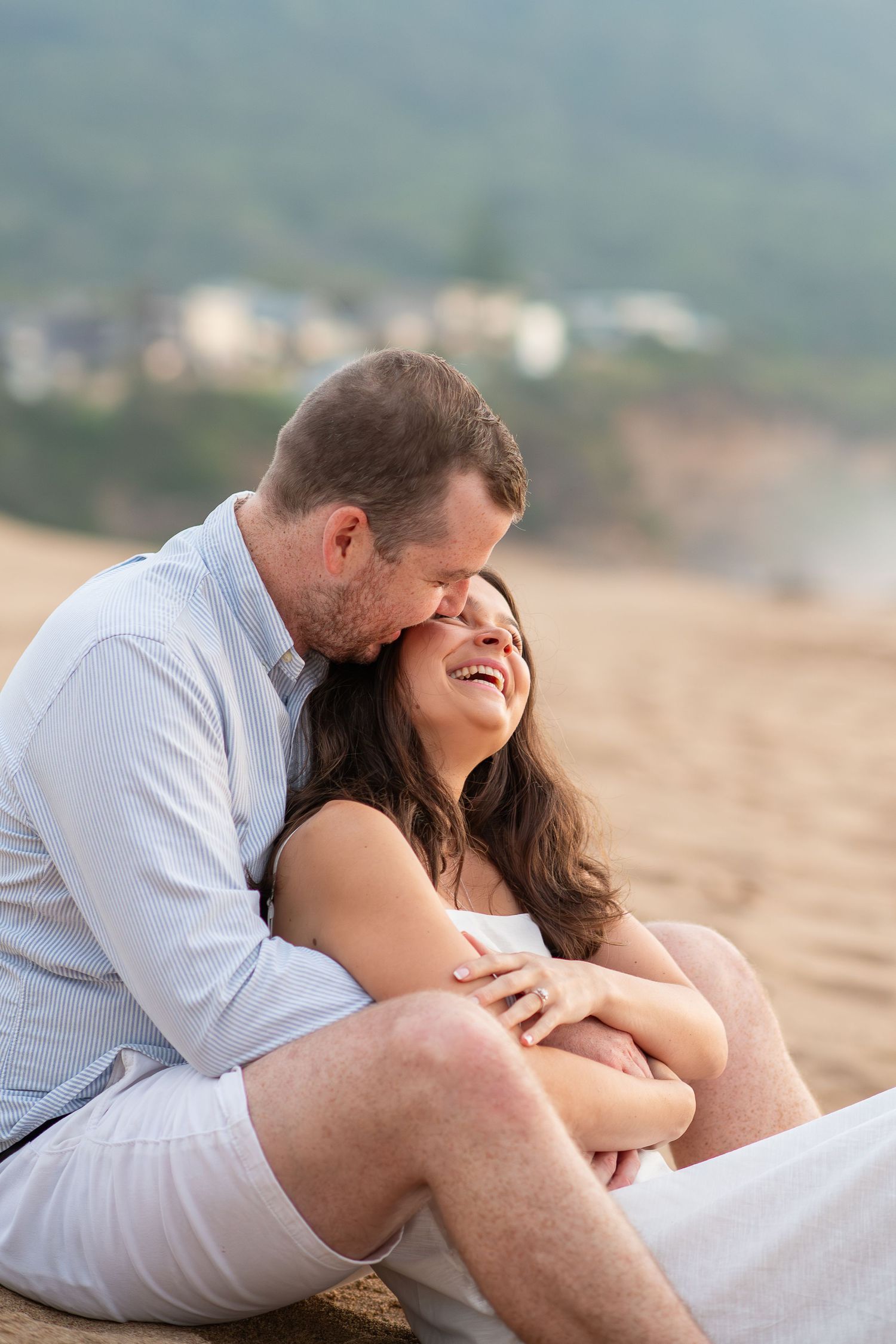Couple sitting on sandy beach, man kissing woman's cheek as she laughs joyfully, with blurred coastal scenery behind them.