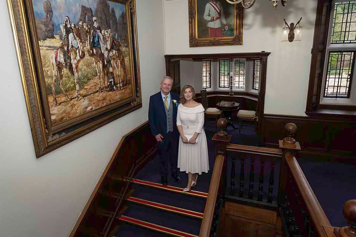 Elegant couple posing on a staircase in a historic building with a large oil painting and wood paneling.