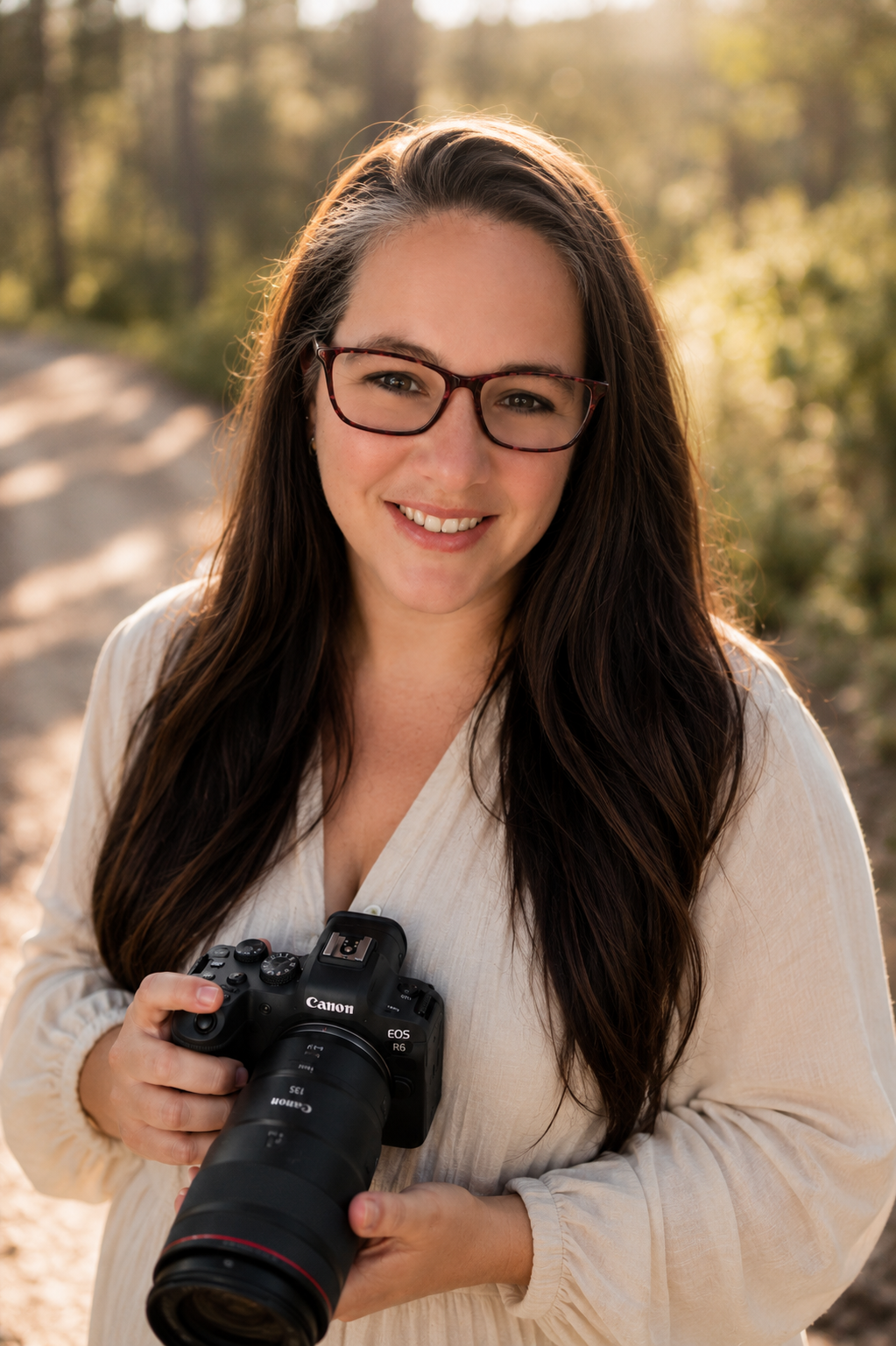 Woman with long dark hair and glasses smiles while holding a Canon EOS camera outdoors in golden sunlight.