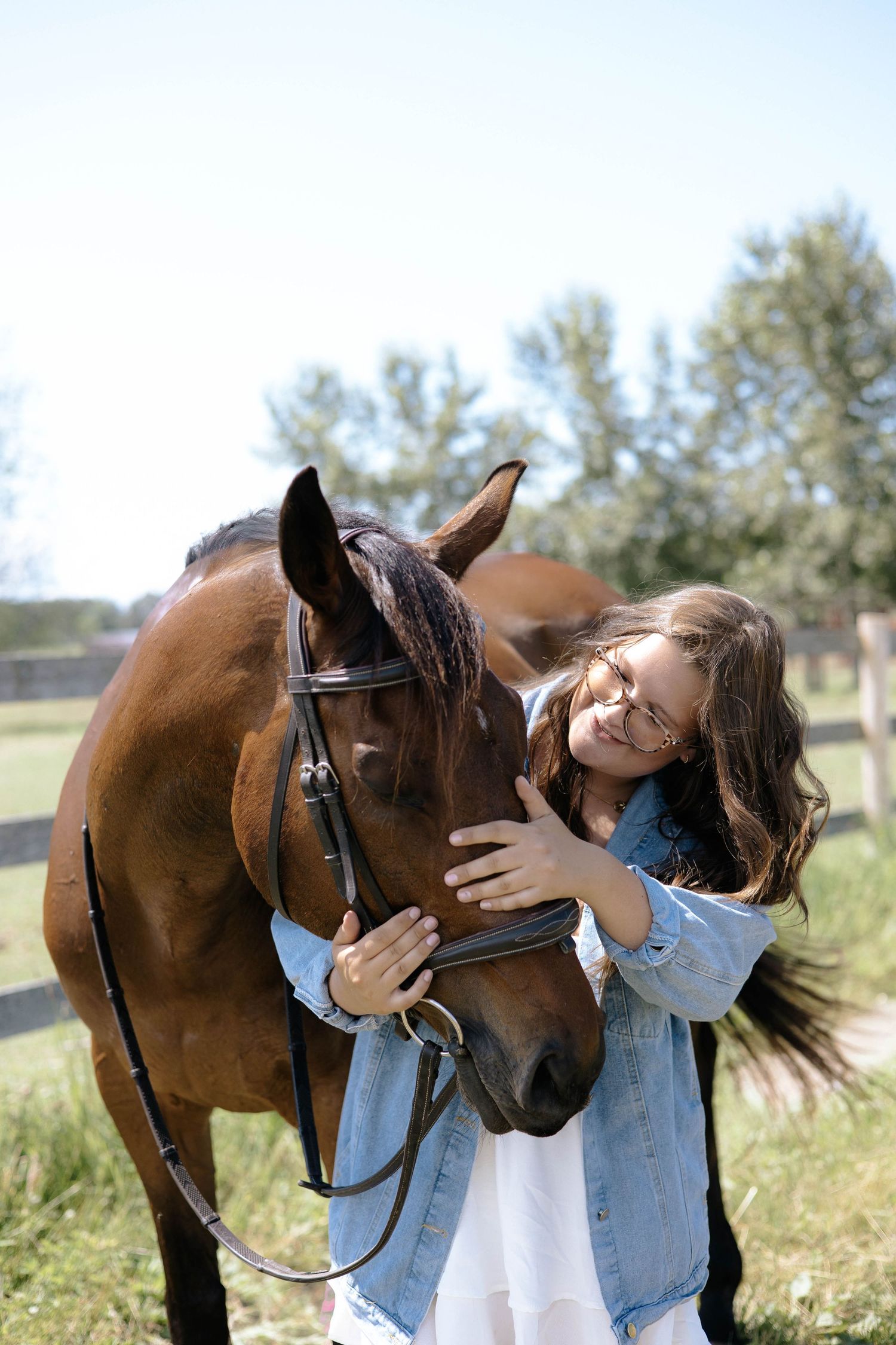 A young woman in a denim jacket affectionately nuzzles a bay horse in a sunny green pasture.