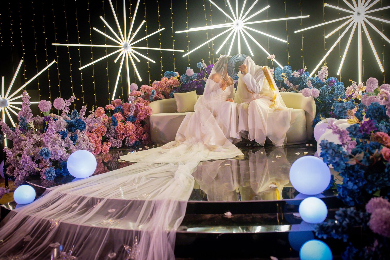 Two brides in white gowns sit surrounded by colorful floral arrangements and starburst lights at a wedding reception.