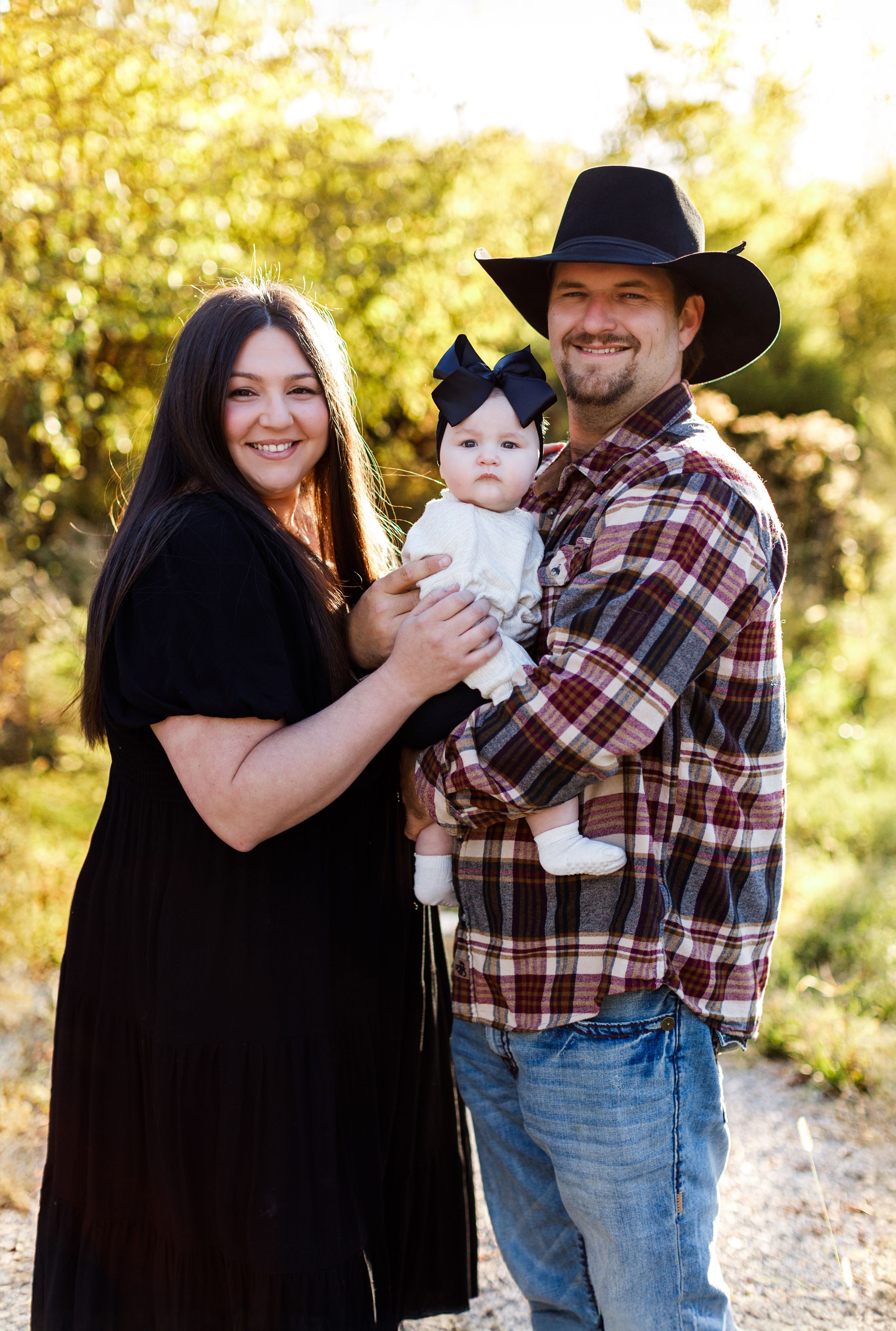 A family poses outdoors in a natural setting with golden sunlight filtering through trees in the background.