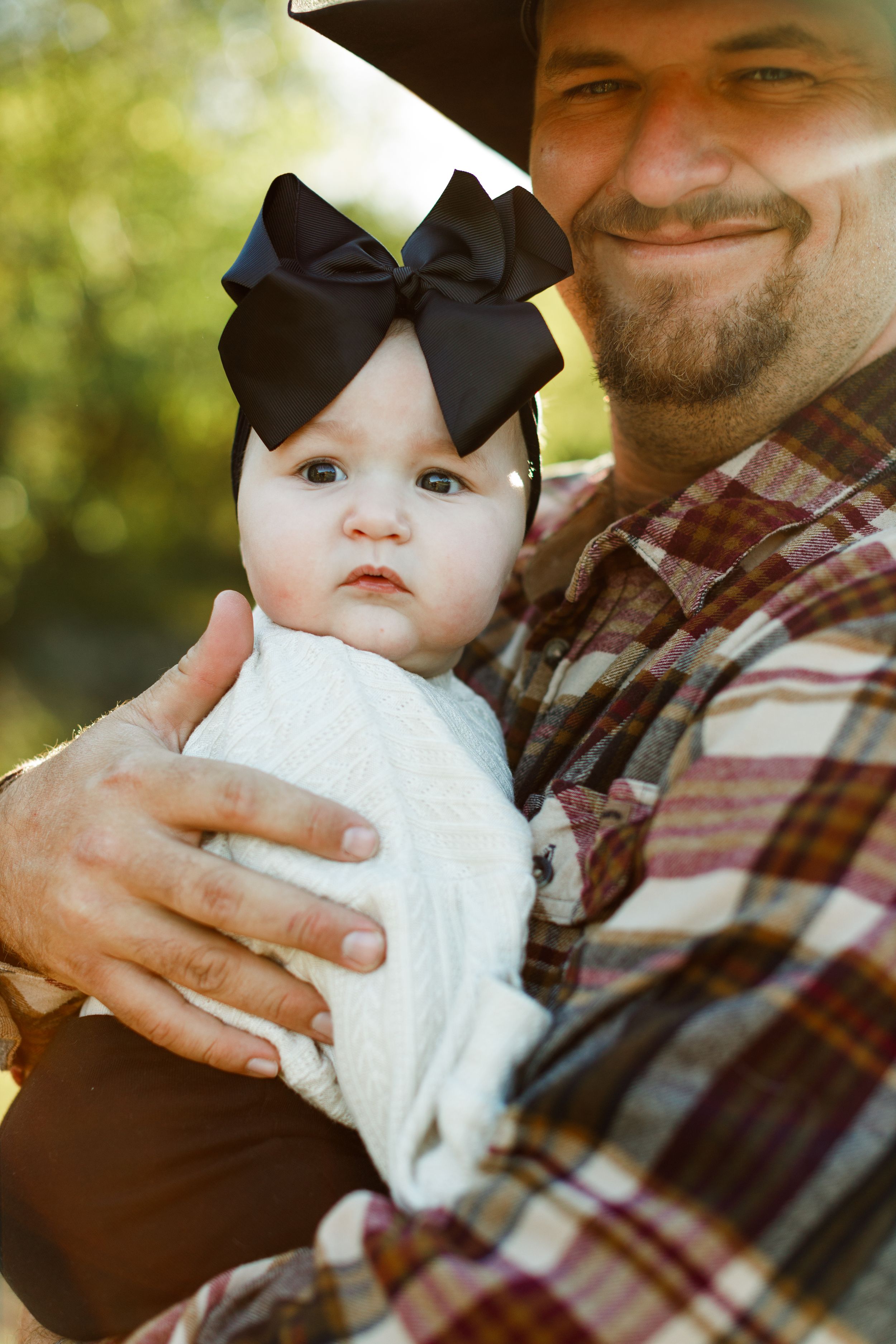 An adorable baby wearing a white sweater and big black bow headband is held lovingly outdoors in the sunshine.