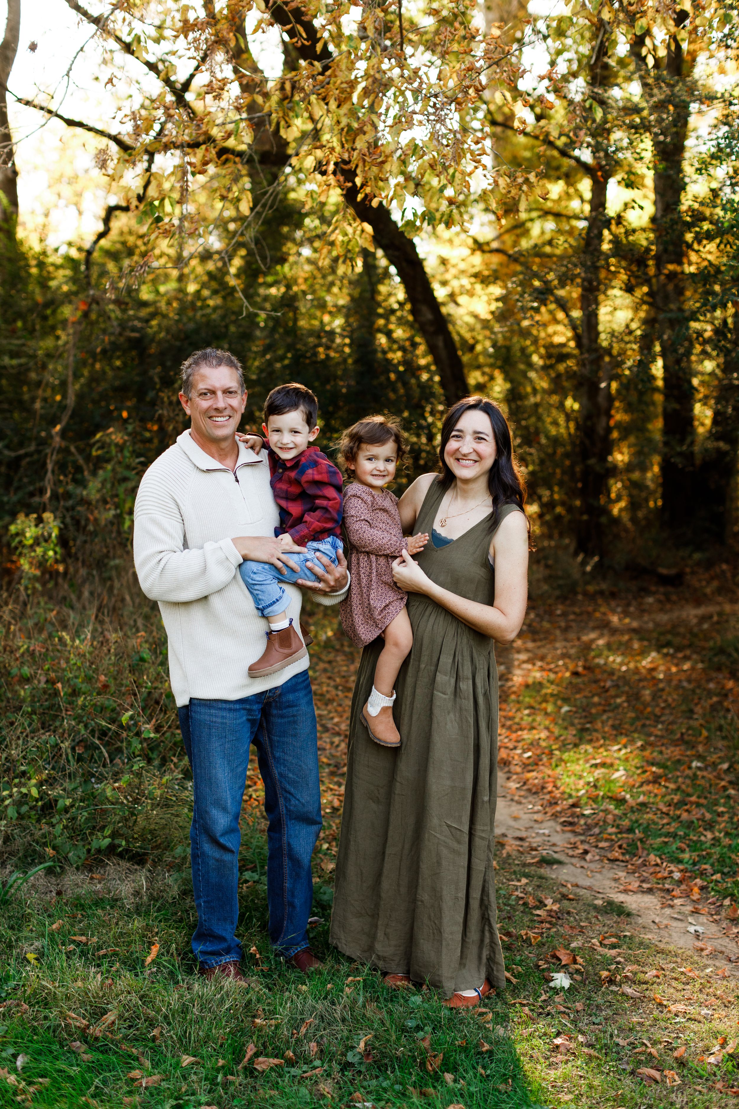 Family portrait taken outdoors on a fall day with golden sunlight filtering through autumn trees on a forest path.