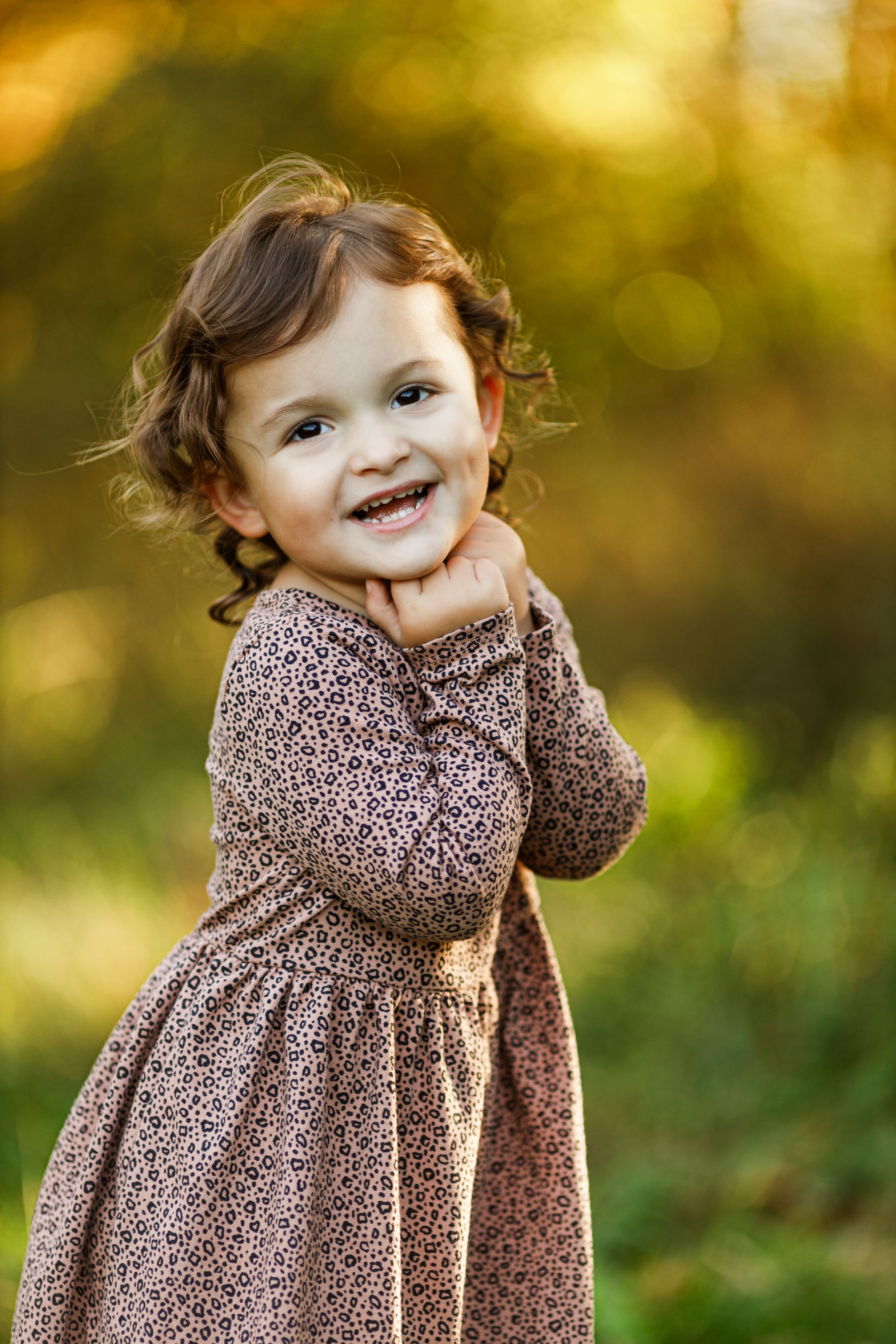 A child wearing a brown polka dot dress poses playfully outdoors with golden bokeh background lighting.
