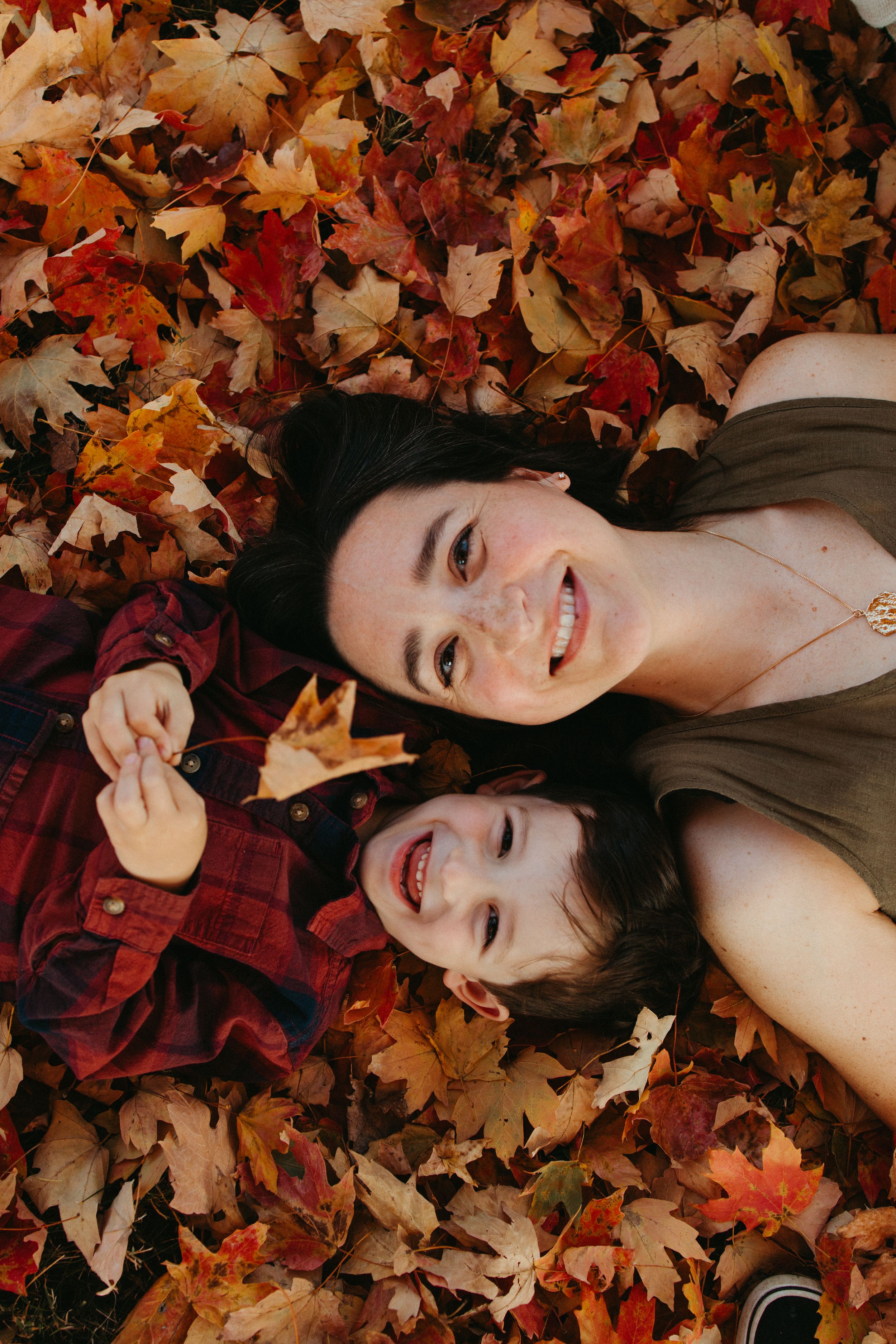 Two people lying in vibrant autumn maple leaves wearing plaid and brown clothing with joyful expressions.