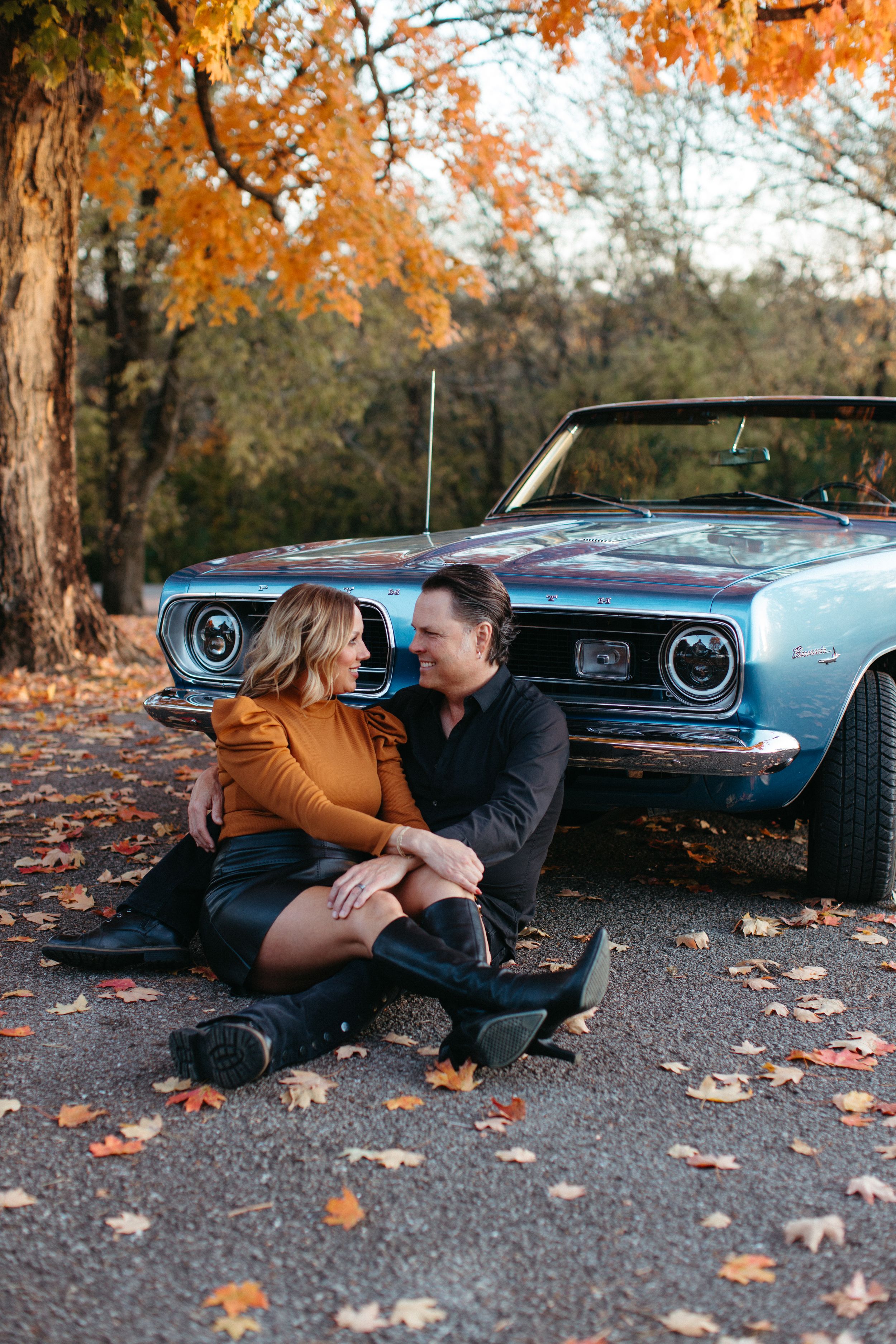 A couple sits together on autumn ground next to a vintage blue convertible car surrounded by orange fall foliage.
