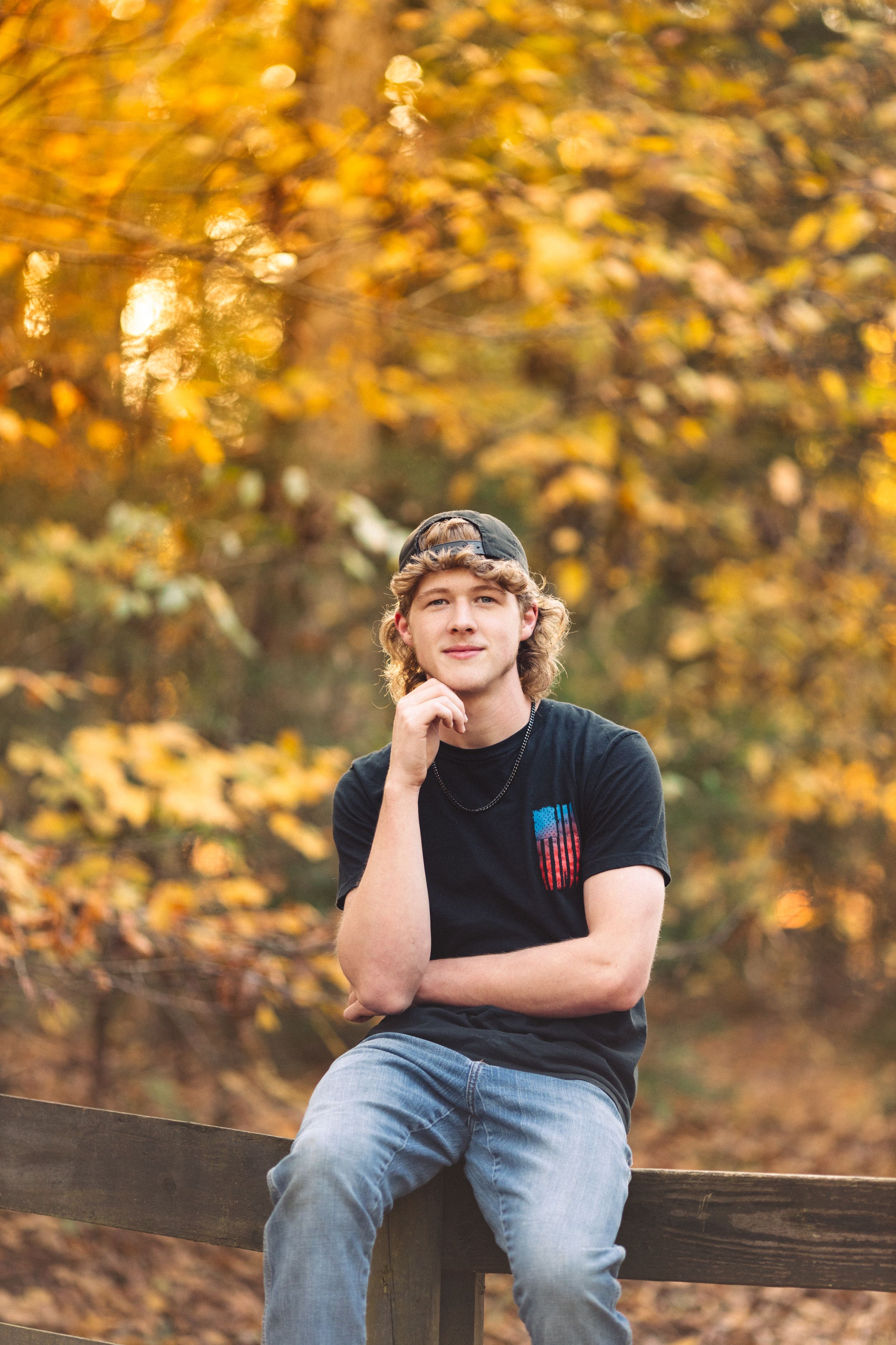 A person sits thoughtfully on a wooden fence wearing a baseball cap and casual attire against golden autumn foliage.