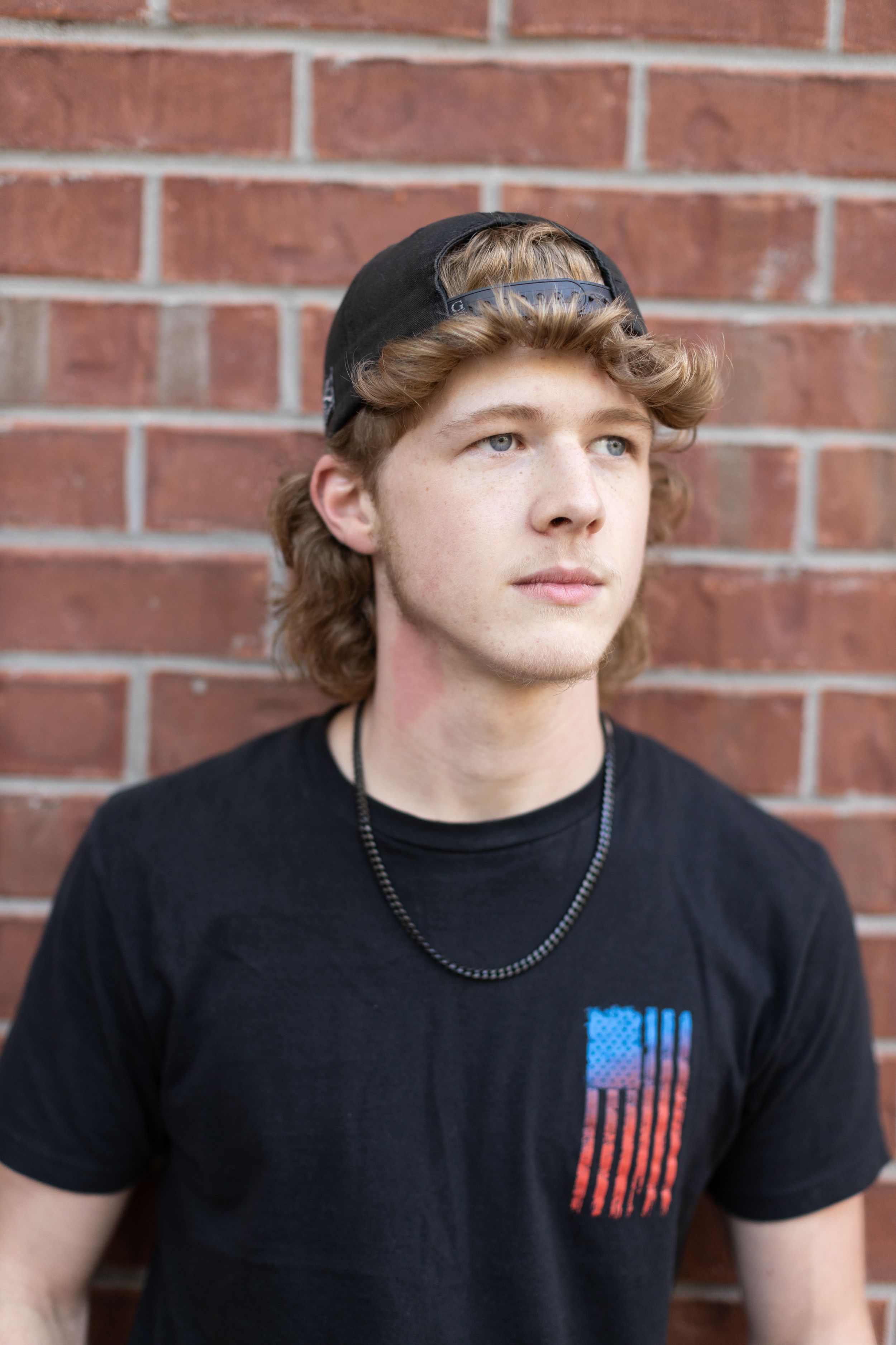 Someone wearing a black t-shirt with an American flag design stands against a red brick wall in a casual pose.