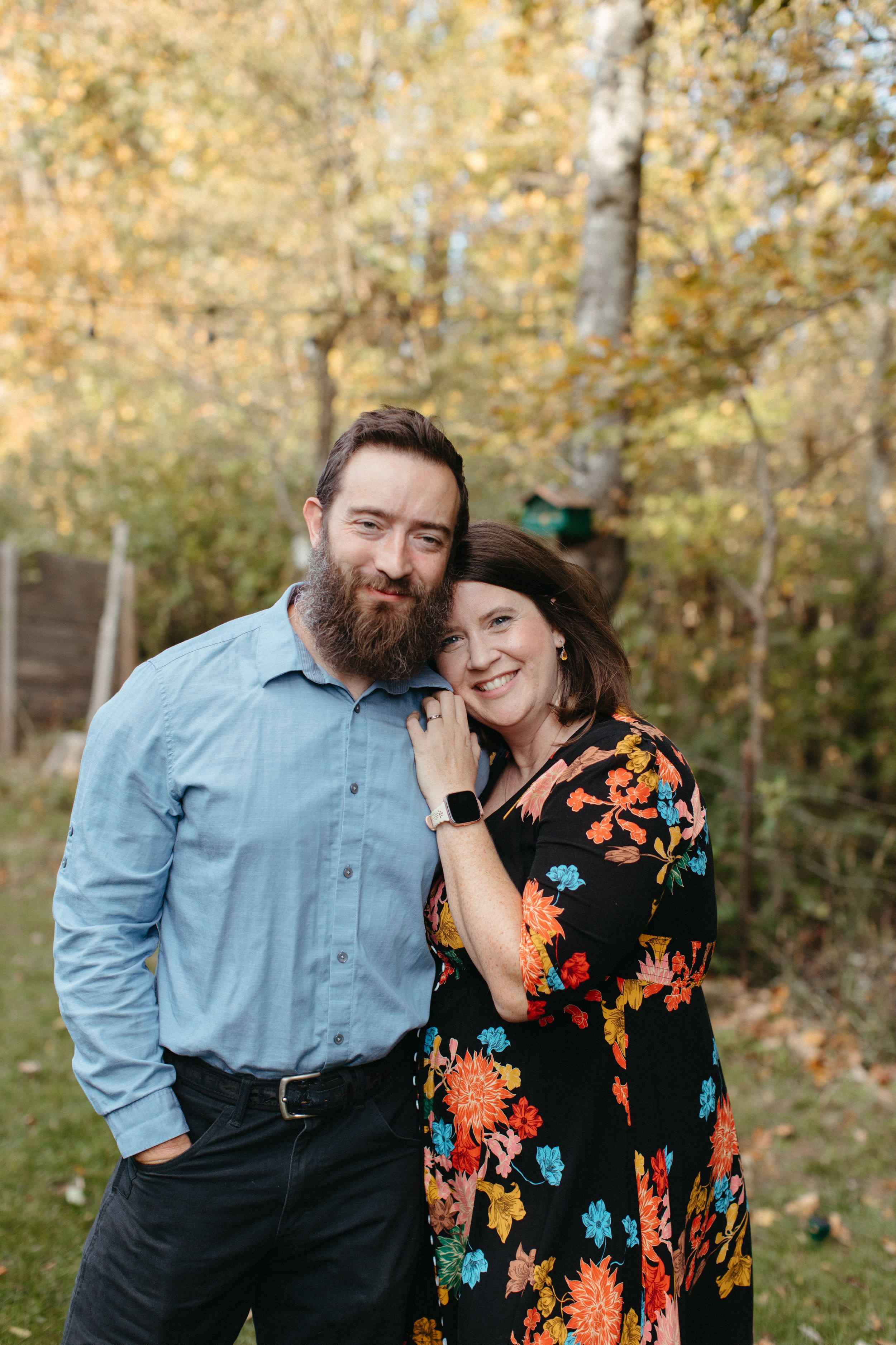 A couple stands together outdoors in autumn, with the person wearing a blue shirt and beard next to someone in a floral dress.