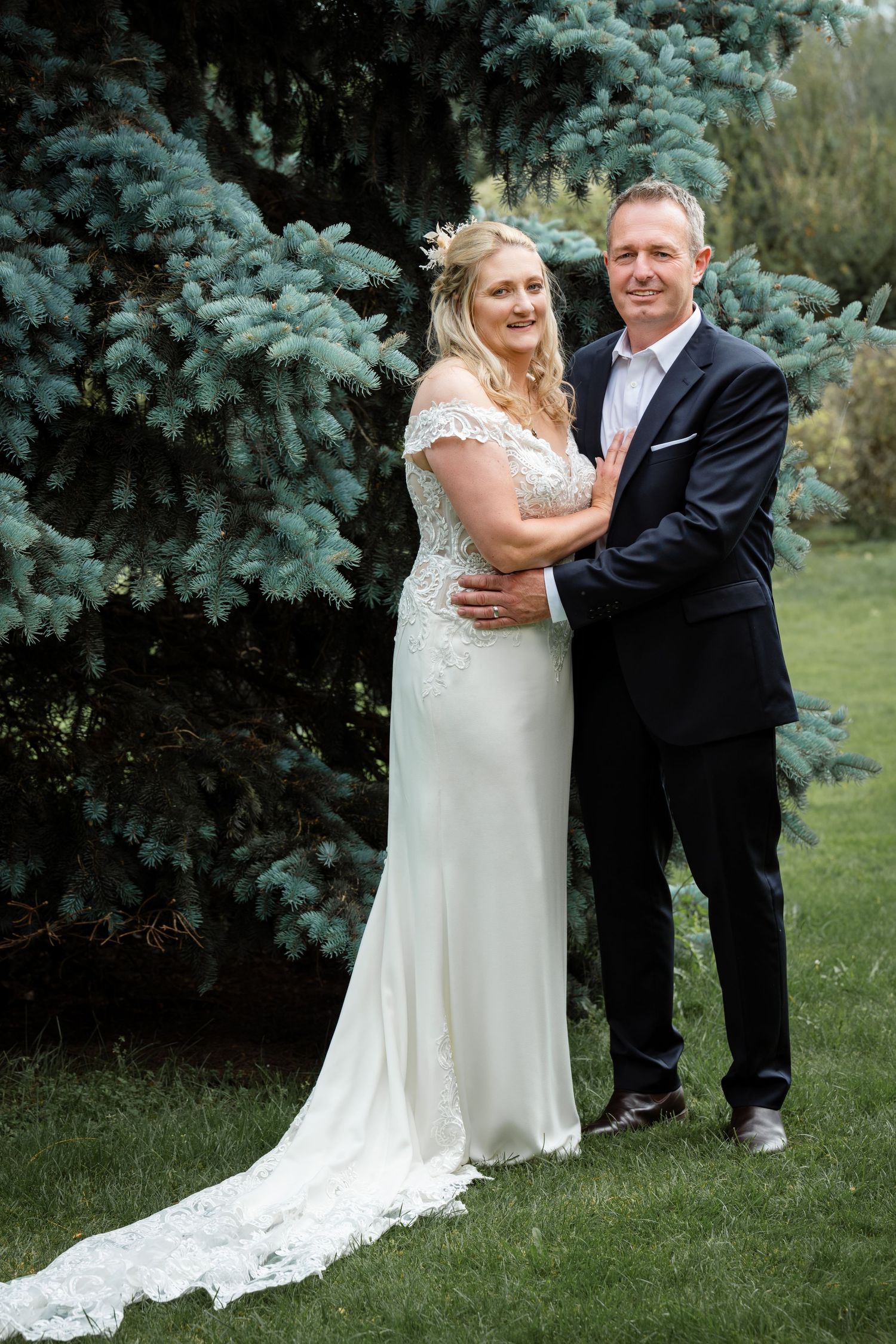 A couple stands together in formal wedding attire in front of blue spruce trees on a lawn.