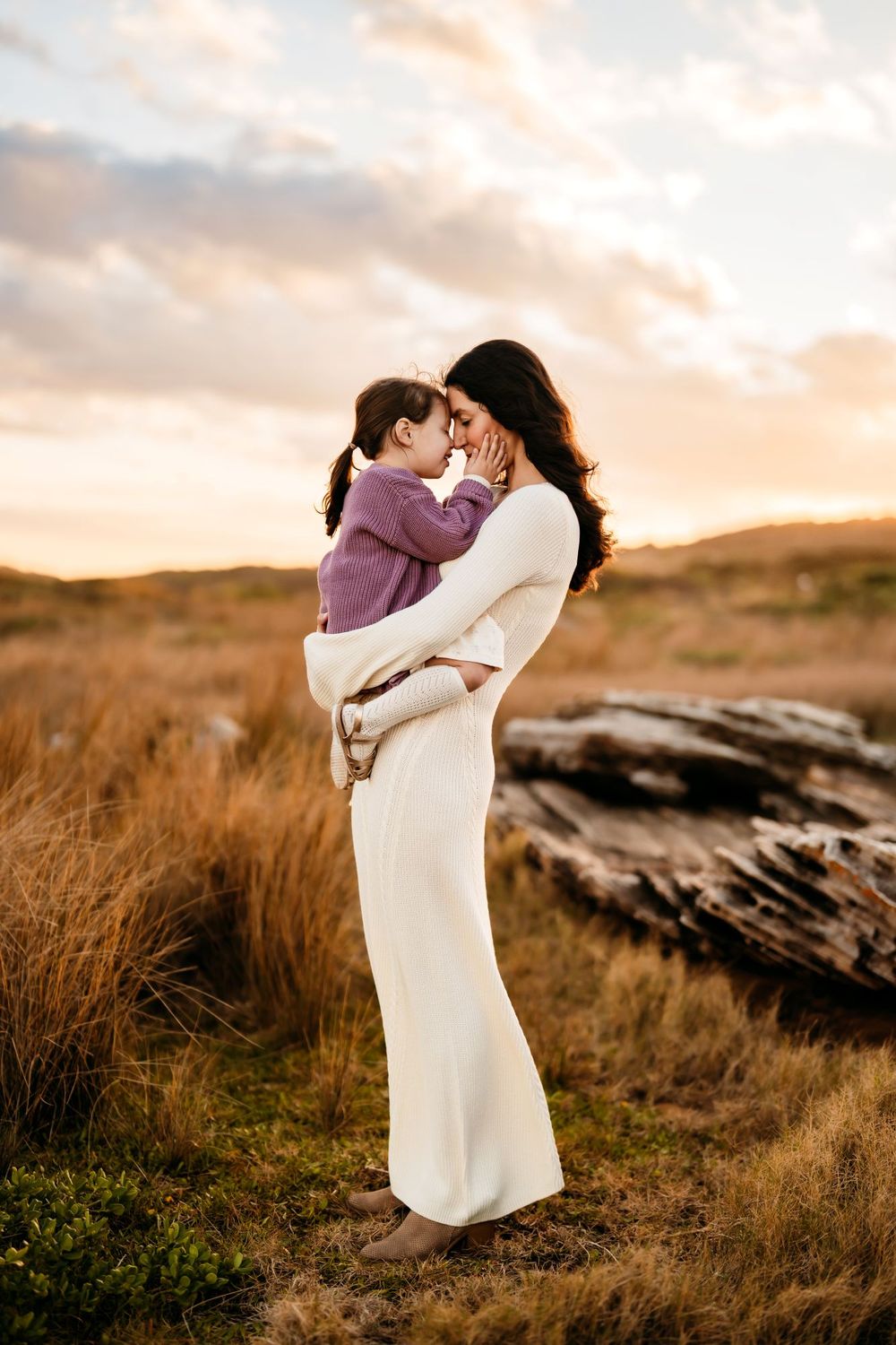 Two people embrace in a white dress against a golden sunset landscape with rocky terrain and tall grass.