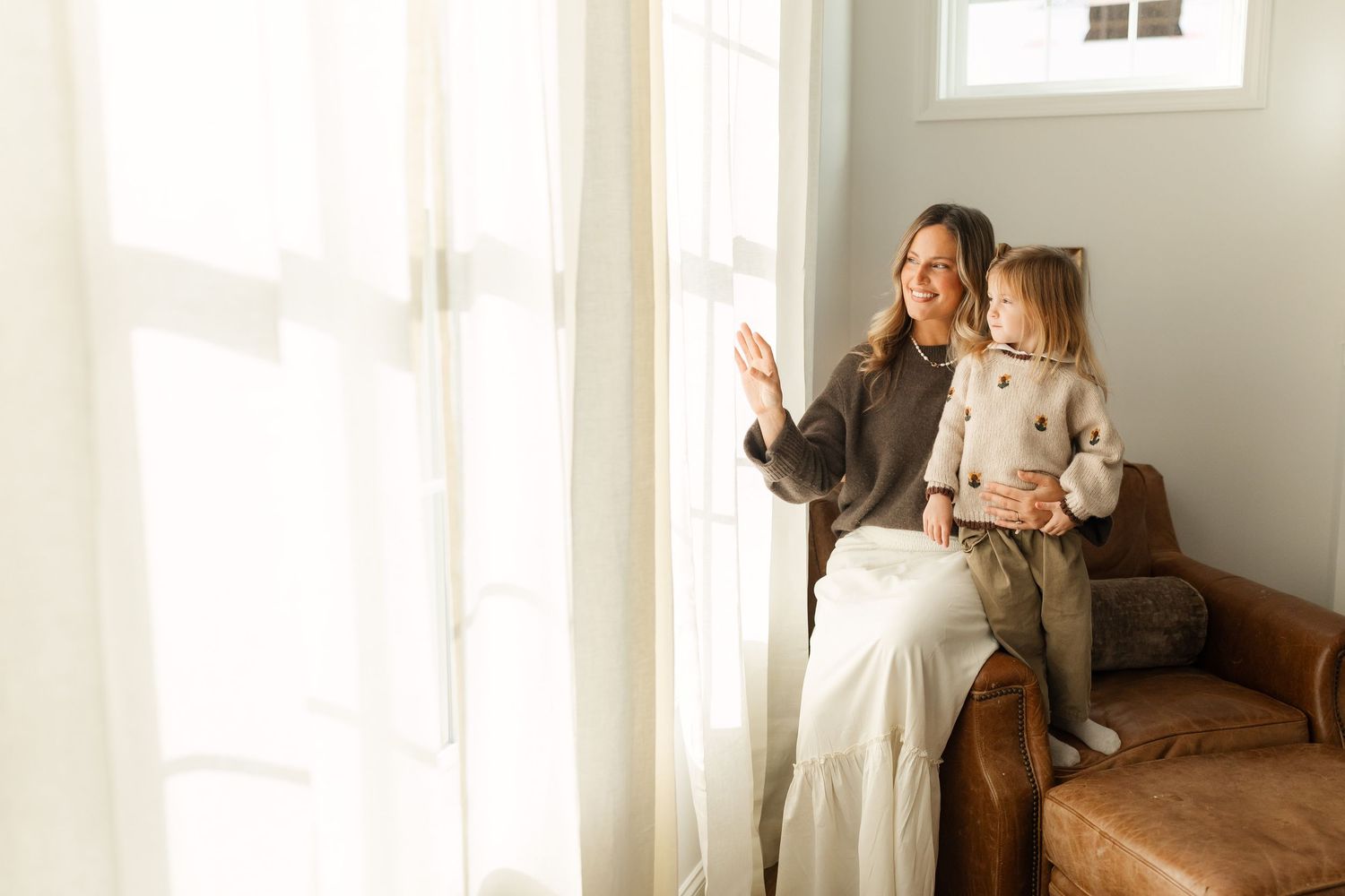 A smiling mother and young daughter sit together on a leather chair by a sunlit window, waving outside.
