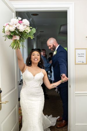 A joyful bride in a fitted lace wedding dress holds up her bouquet of pink and white flowers after her wedding ceremony