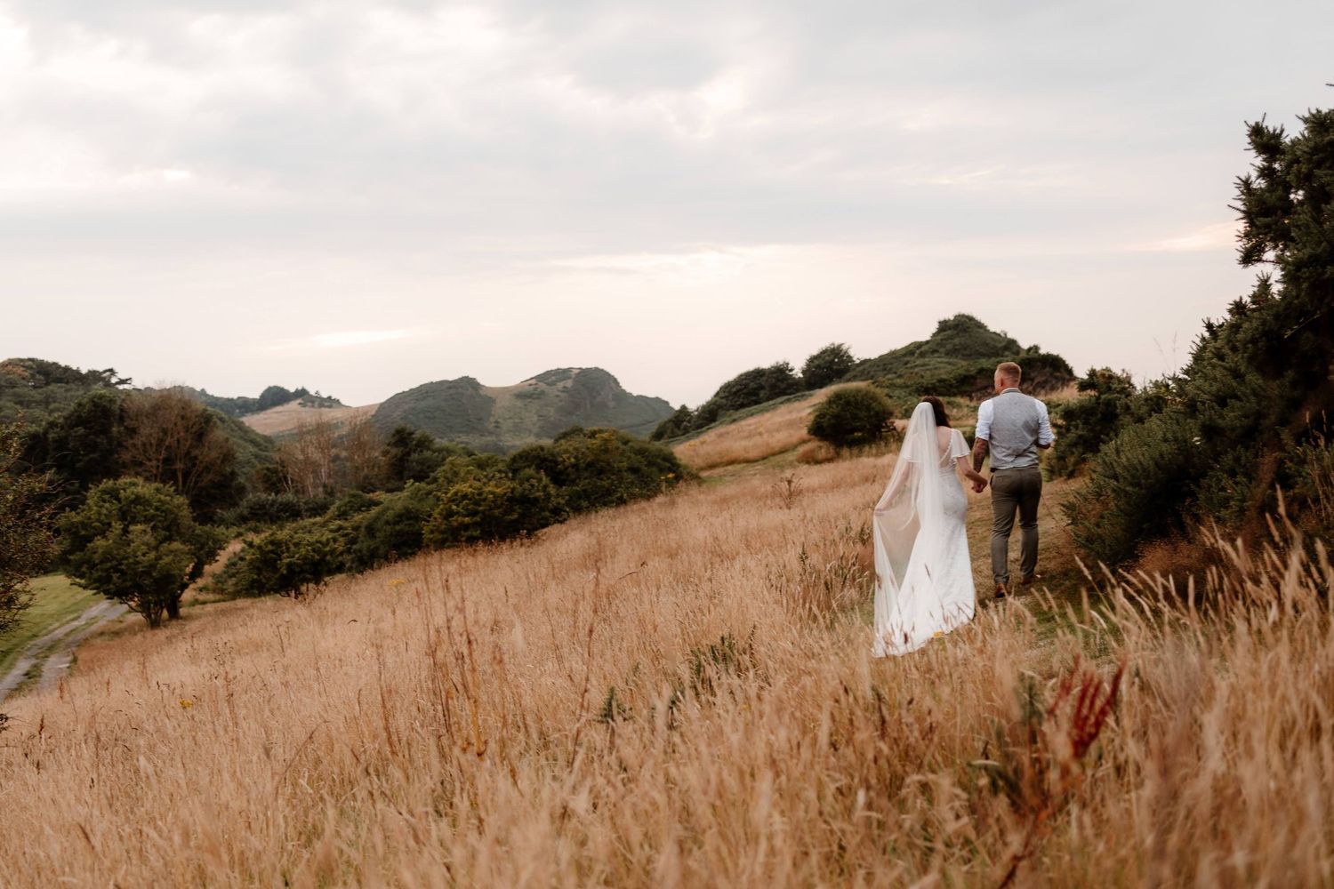 A bride and groom walk hand in hand through the long golden grass at Watermouth Cove wedding venue in North Devon.