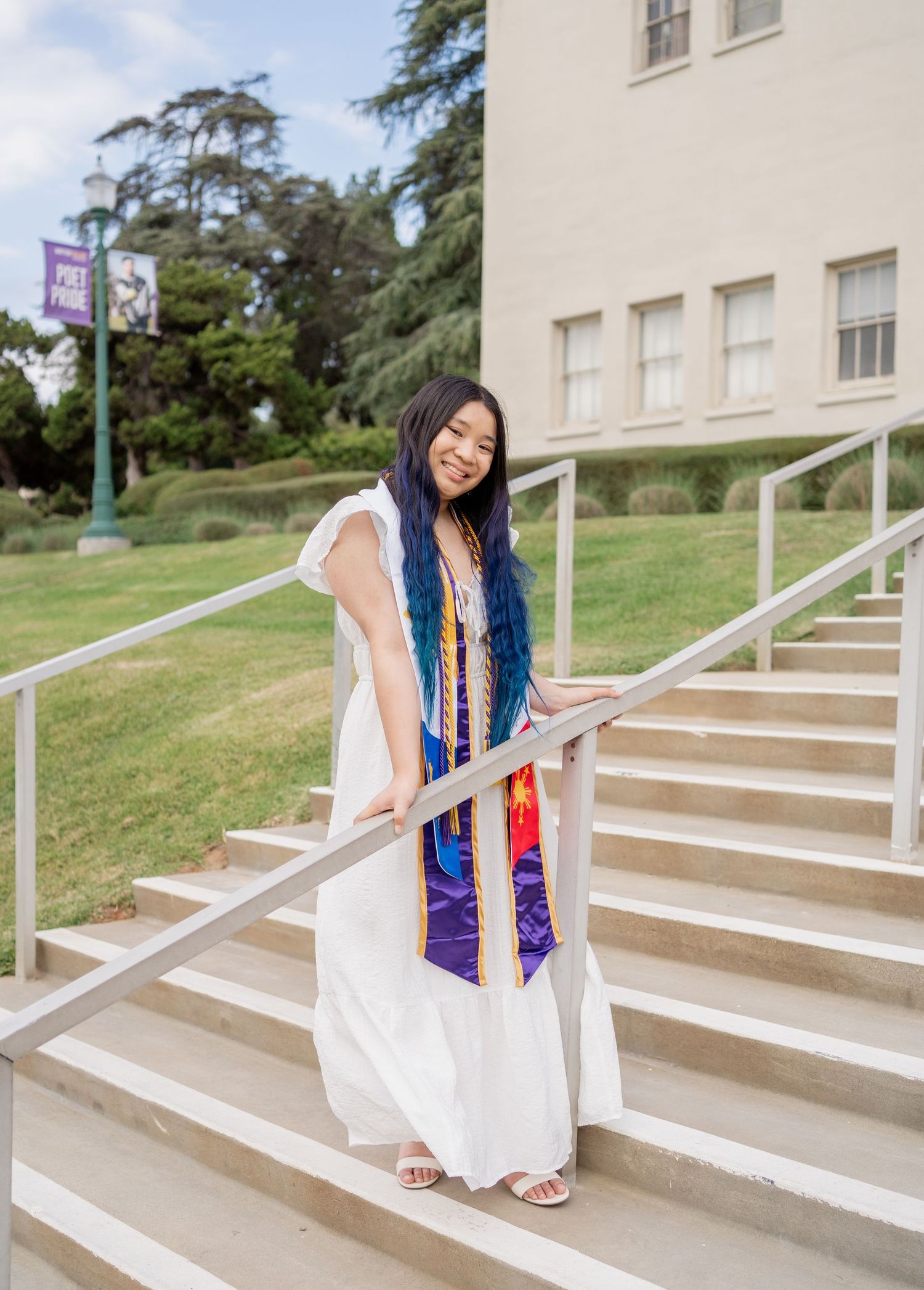 Smiling graduate in white dress and purple honor stole posing on campus stairs at graduation.