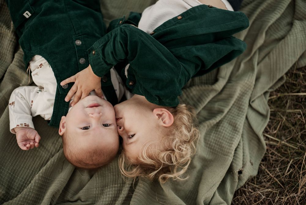 Two young children lying on a green blanket outdoors, a toddler kissing a baby sibling.