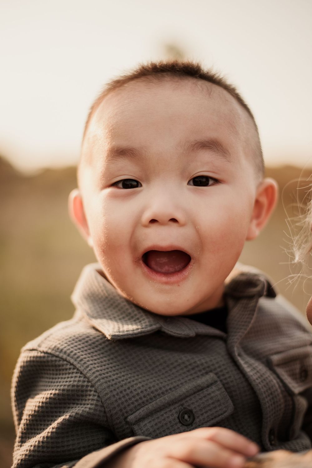 Smiling toddler in a gray jacket stands outdoors during a golden-hour family photo session.