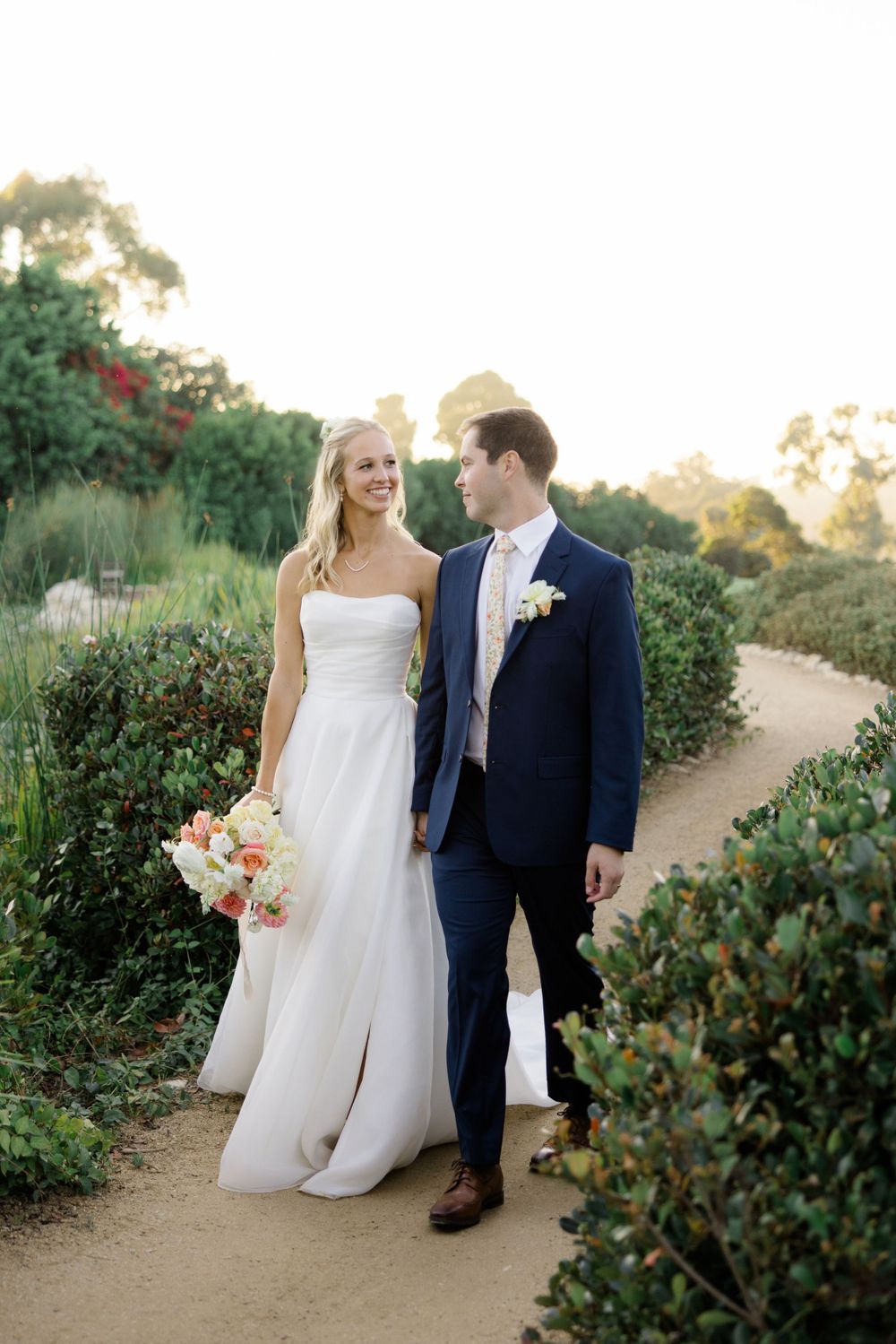 A couple walks along a garden path, with the bride in white and groom in navy during their wedding photoshoot.