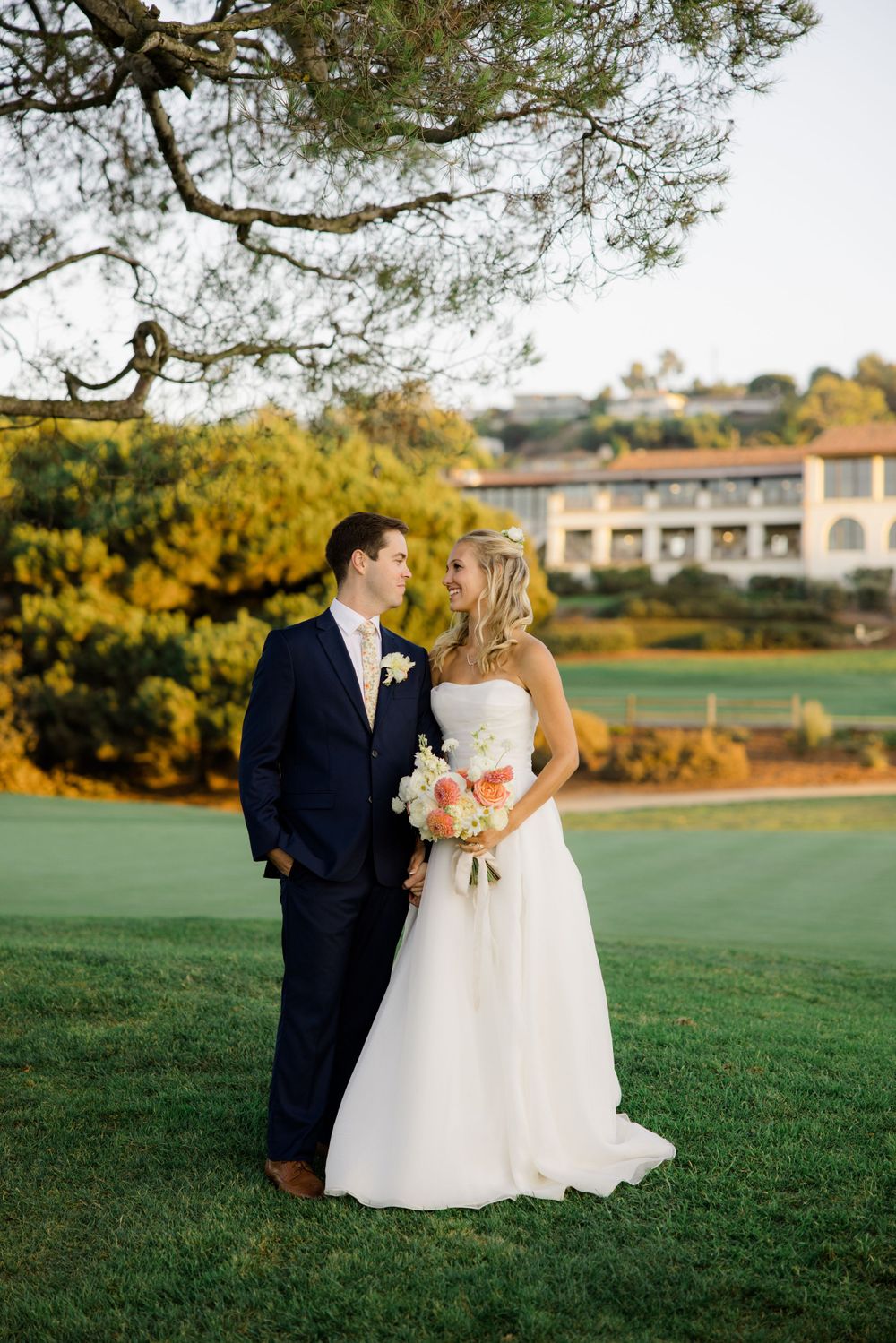 A wedding couple poses on a manicured lawn with a resort building visible in the background at golden hour.