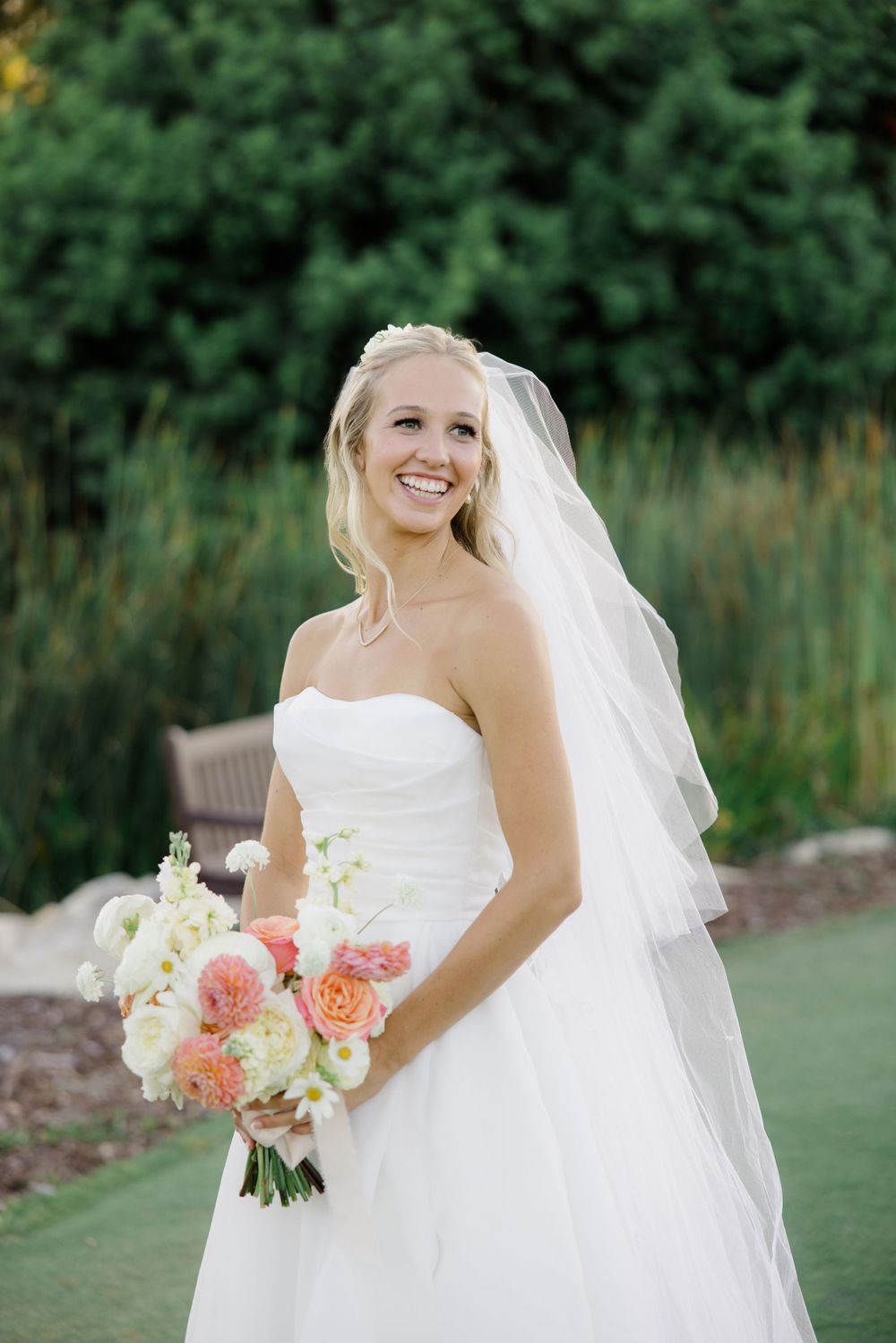 Elegant bride in a white strapless gown holding a coral and white bouquet in a garden setting.