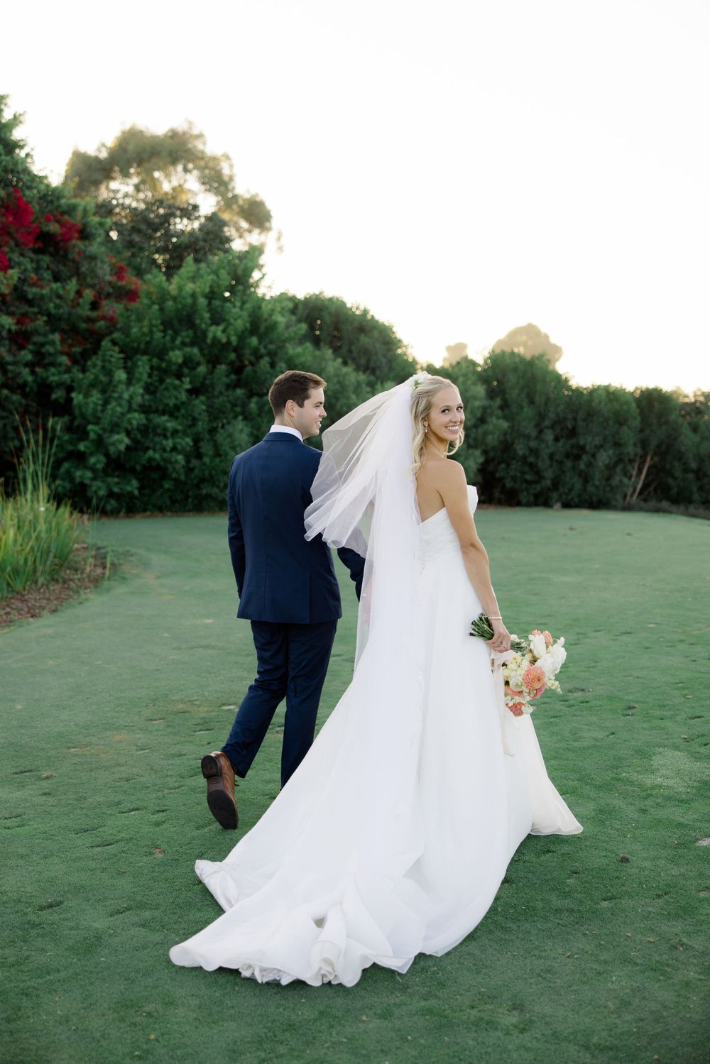 Wedding couple walking across a lush green lawn during golden hour photography session.