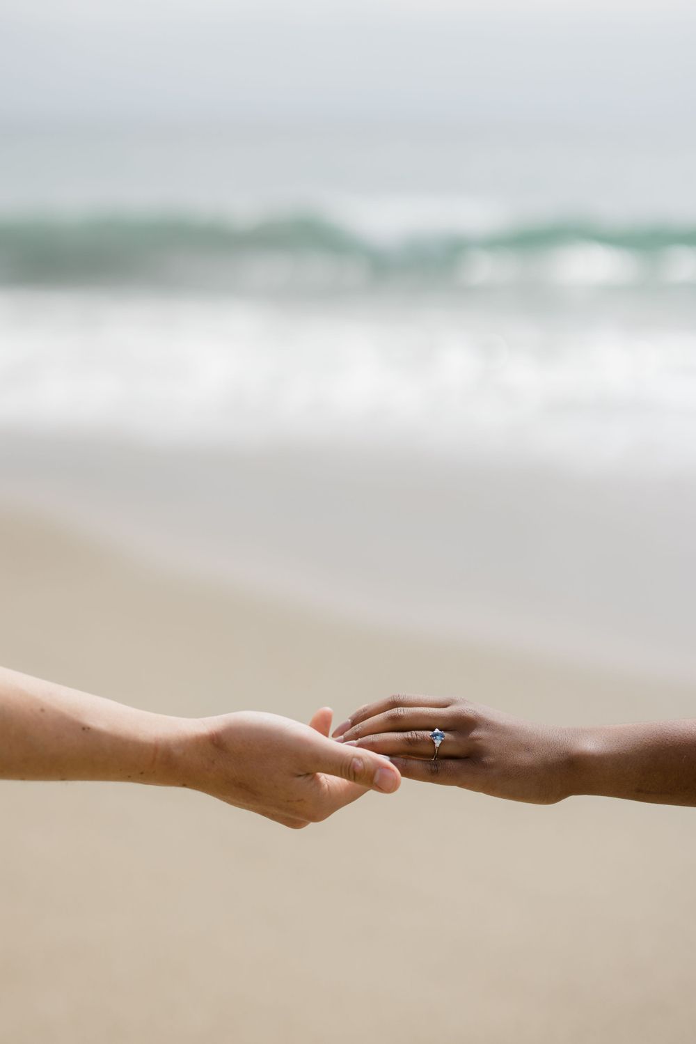 Two hands reach out to touch, showing an engagement ring against a beach backdrop.