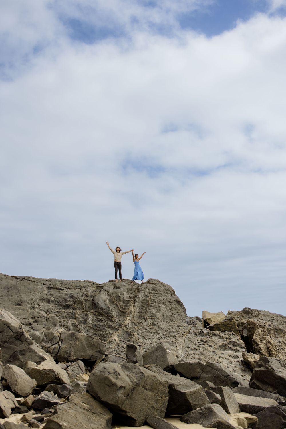 Two people stand triumphantly on a rocky cliff overlooking the ocean.