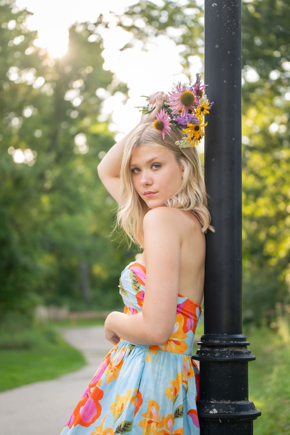 Young blonde woman in floral dress and flower crown leans against black lamp post in sunny park.