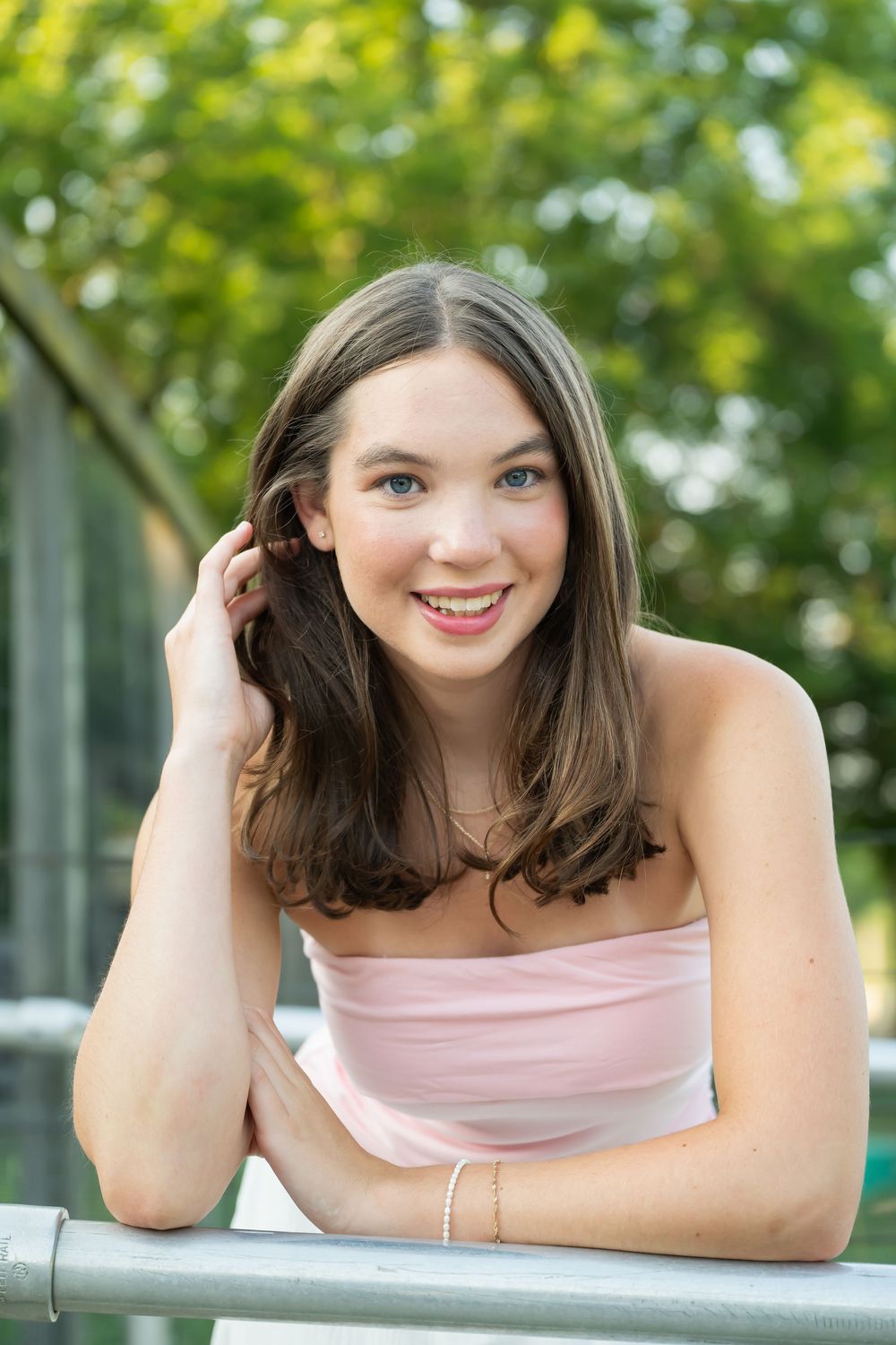 Young woman in pink strapless top smiling outdoors, leaning on a railing with green trees in background.