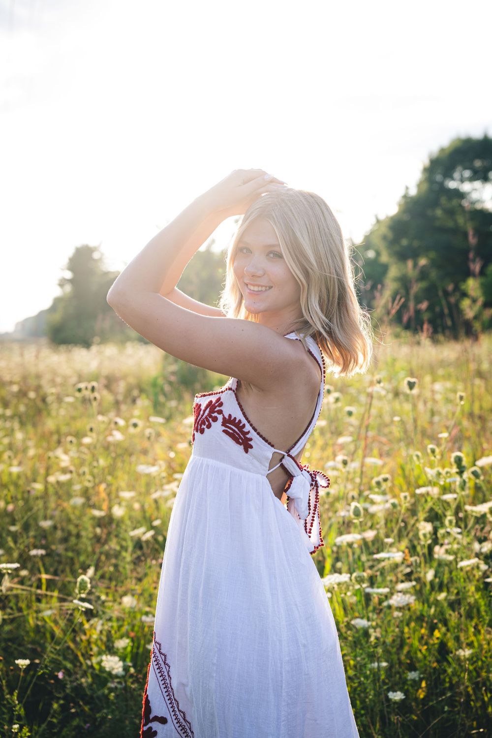 Young blonde woman in white embroidered backless dress smiling in a sunlit wildflower meadow.
